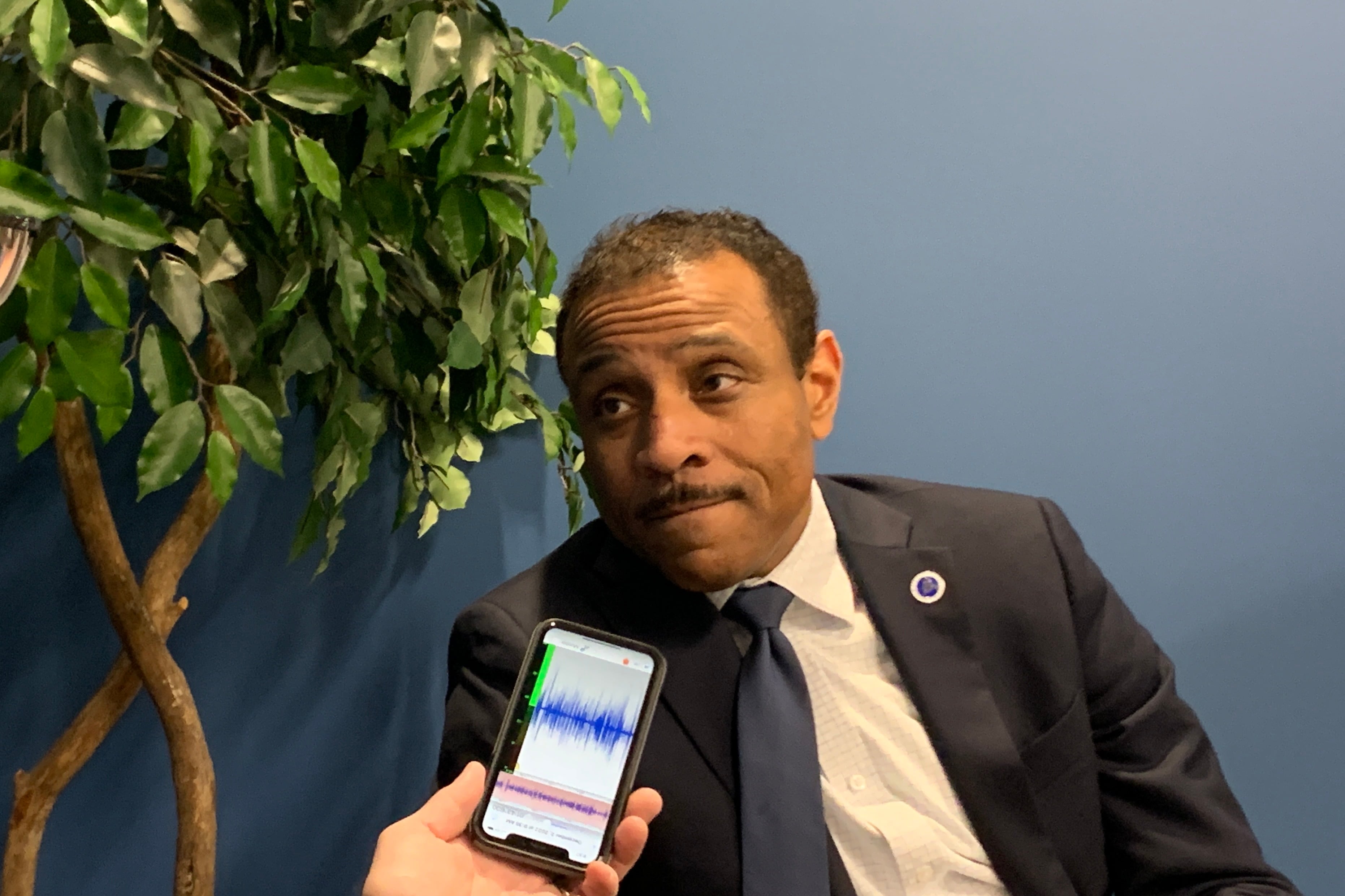 A man in a dark suit and white shirt leans to his right while a person holds a phone near his face in front of a plant a blue wall.