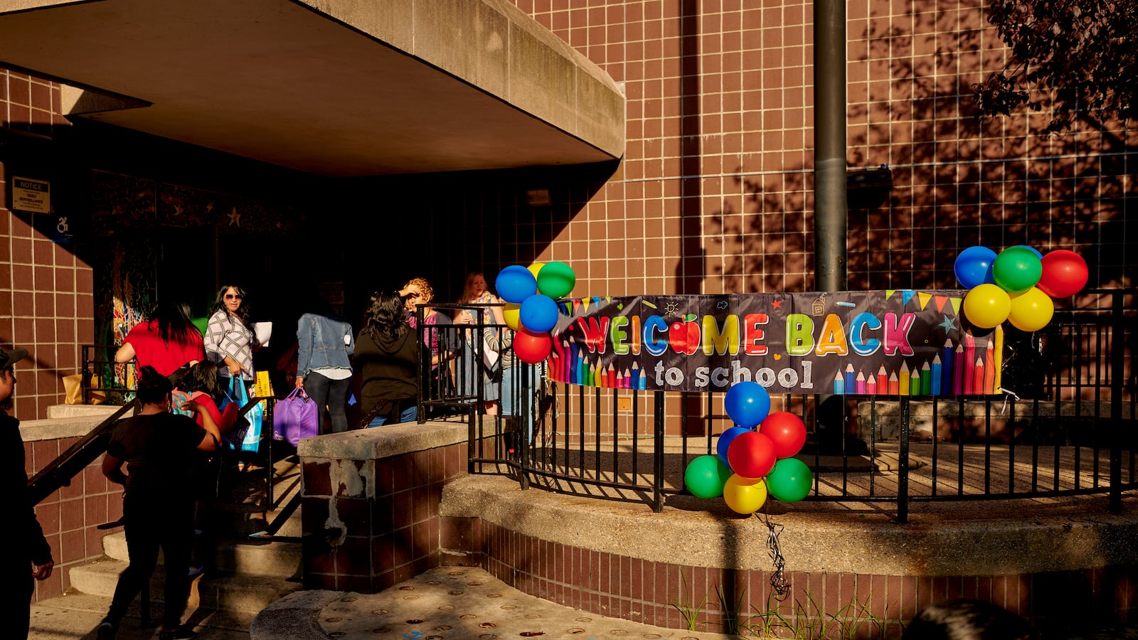 A sign reads “Welcome Back to School” outside of a large brick building as students walk into its doors.