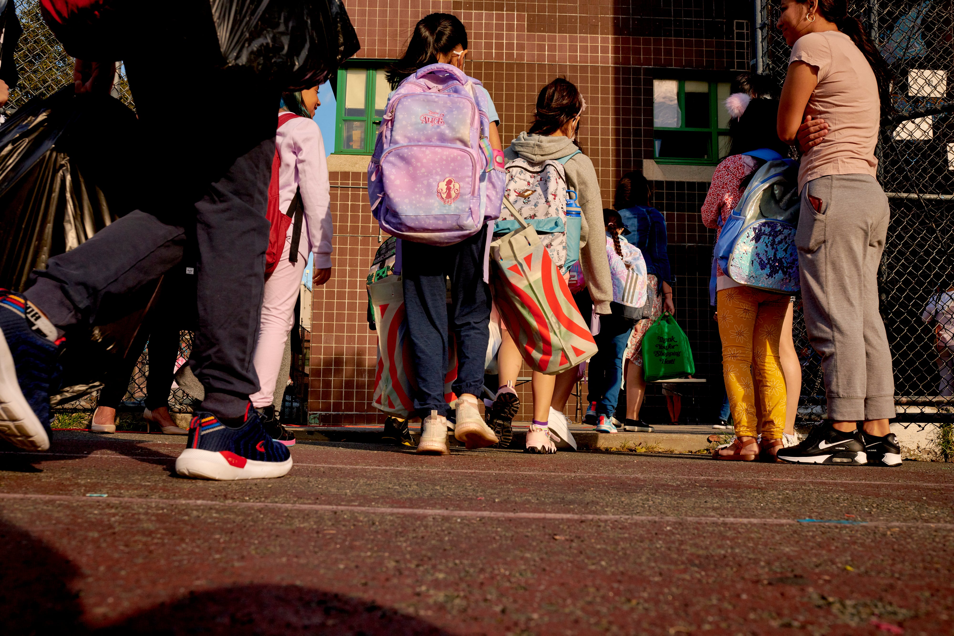 A group of young students wearing backpacks line up in front of a large brick building.