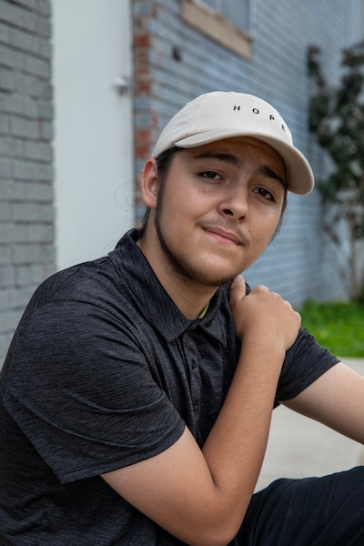 A young man wearing a tan baseball cap and a grey shirt poses for a portrait outside of a building.