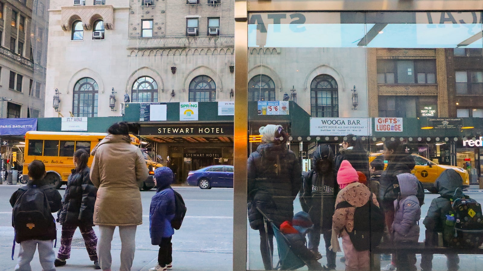 A group of adults and young children stand by a bus stop on a busy city street.