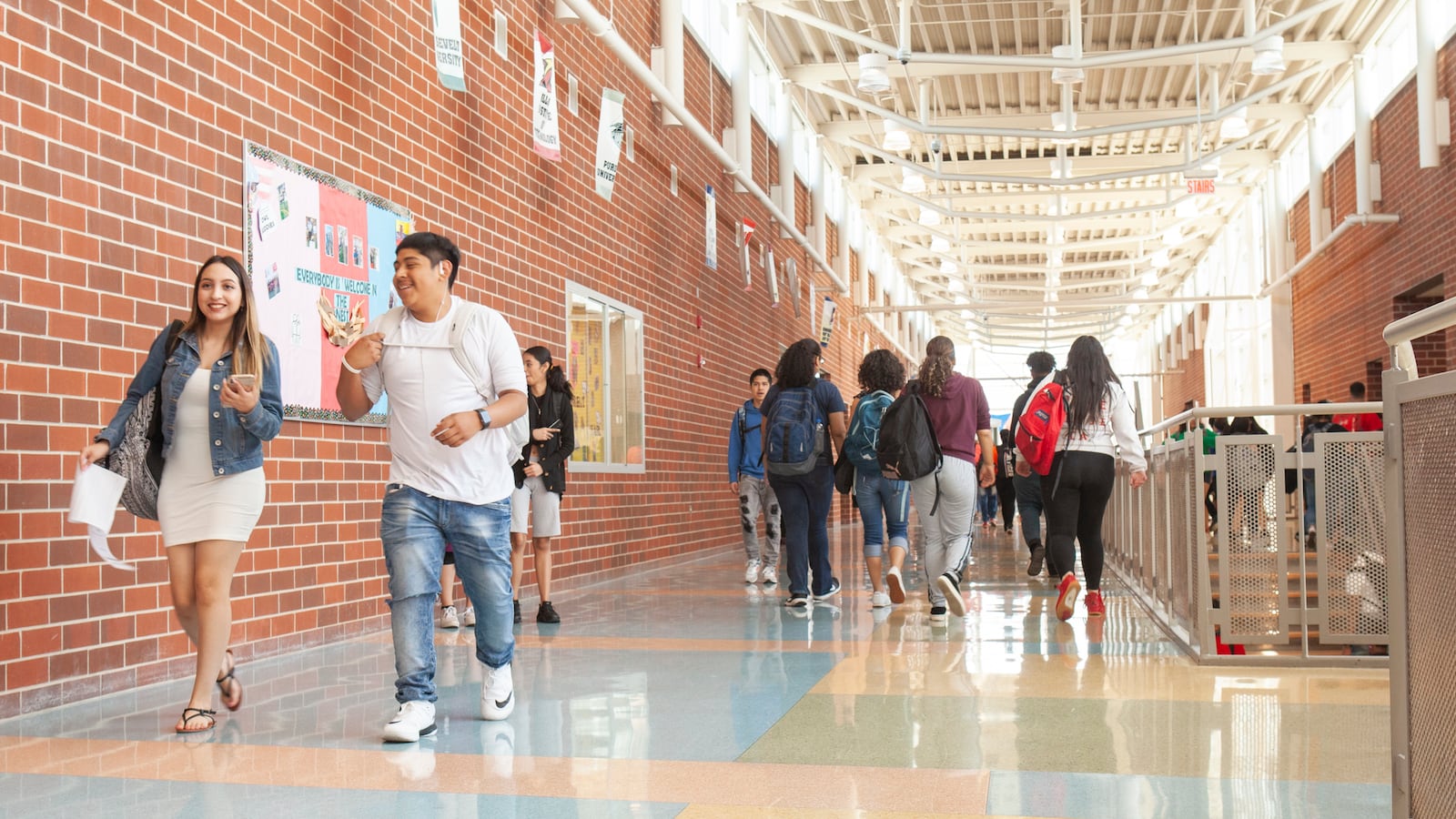Students walk in a hallway at a school. There is a red brick wall on the left with a high ceiling and light shining through.