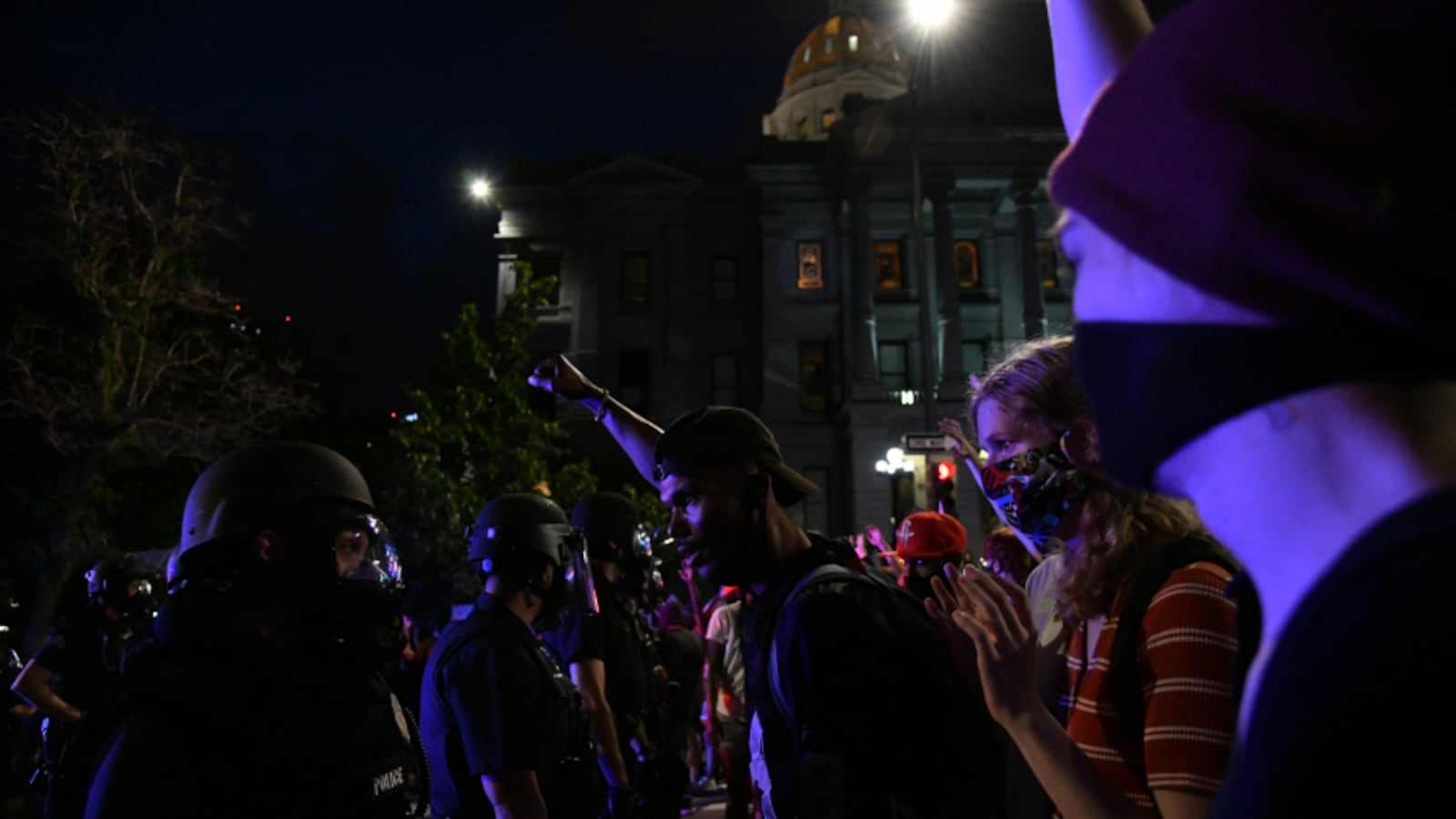 Demonstrators protesting the death of George Floyd at the hands of Minneapolis police face off with Denver Police officers outside the Colorado Capitol. A blue light highlights the faces of all on the scene.