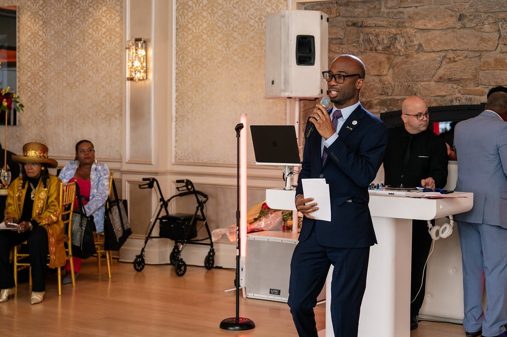 A man in a suit and glasses holds a mic and stands in front of a podium.