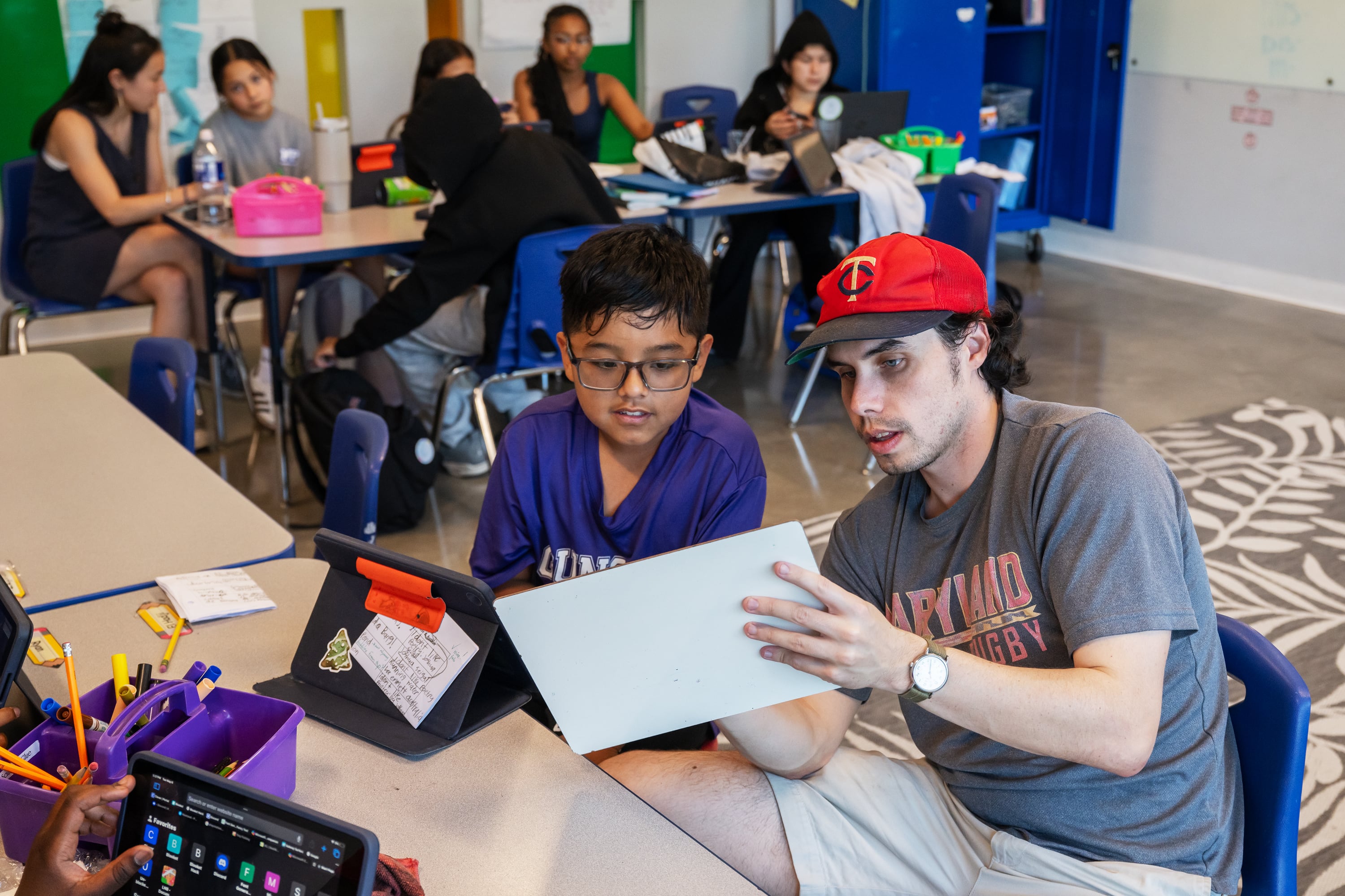 A man in a gray t-shirt and red cap shows a paper to a boy in glasses and a purple t-shirt.