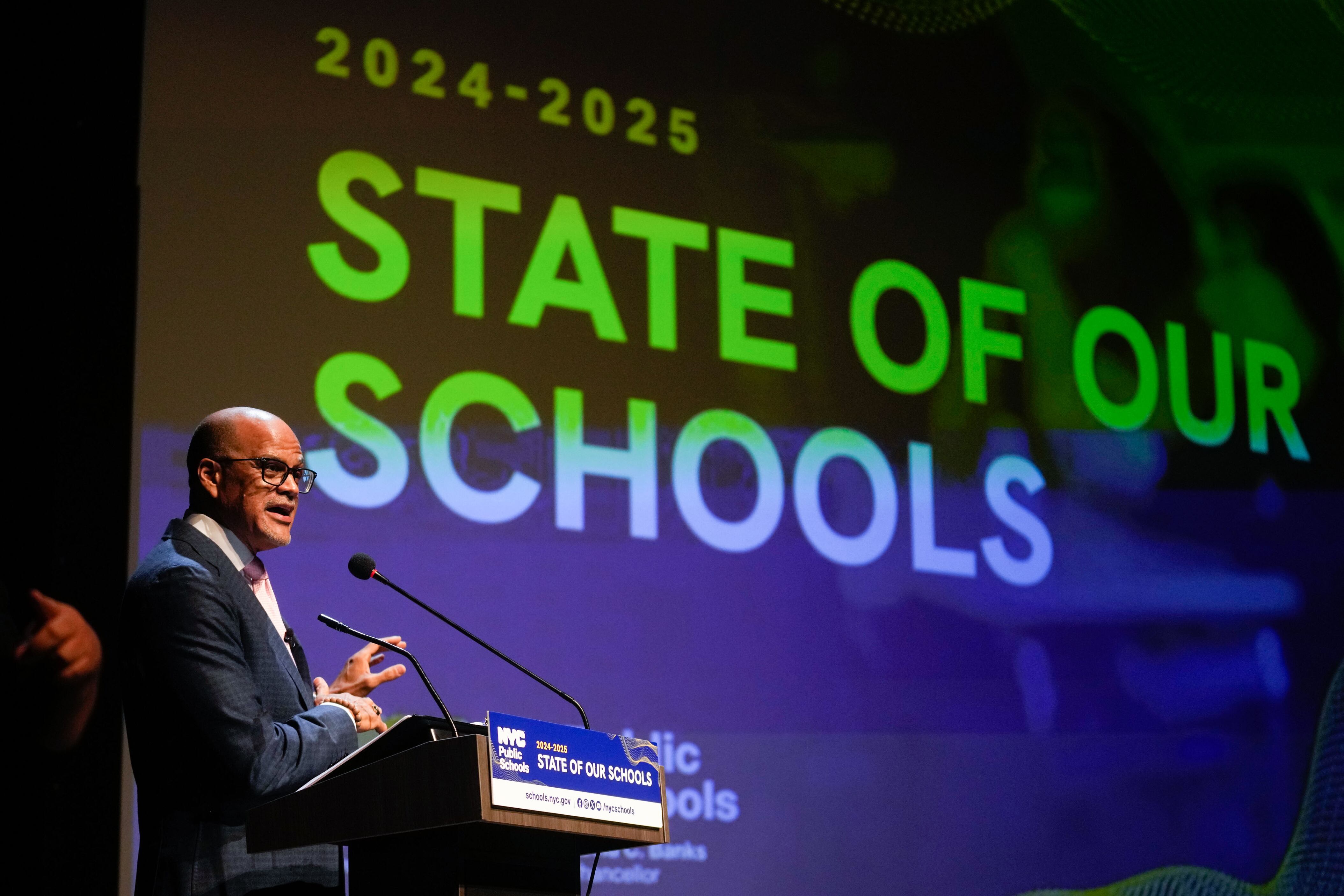 A man in glasses and a suit stands in front of a screen.