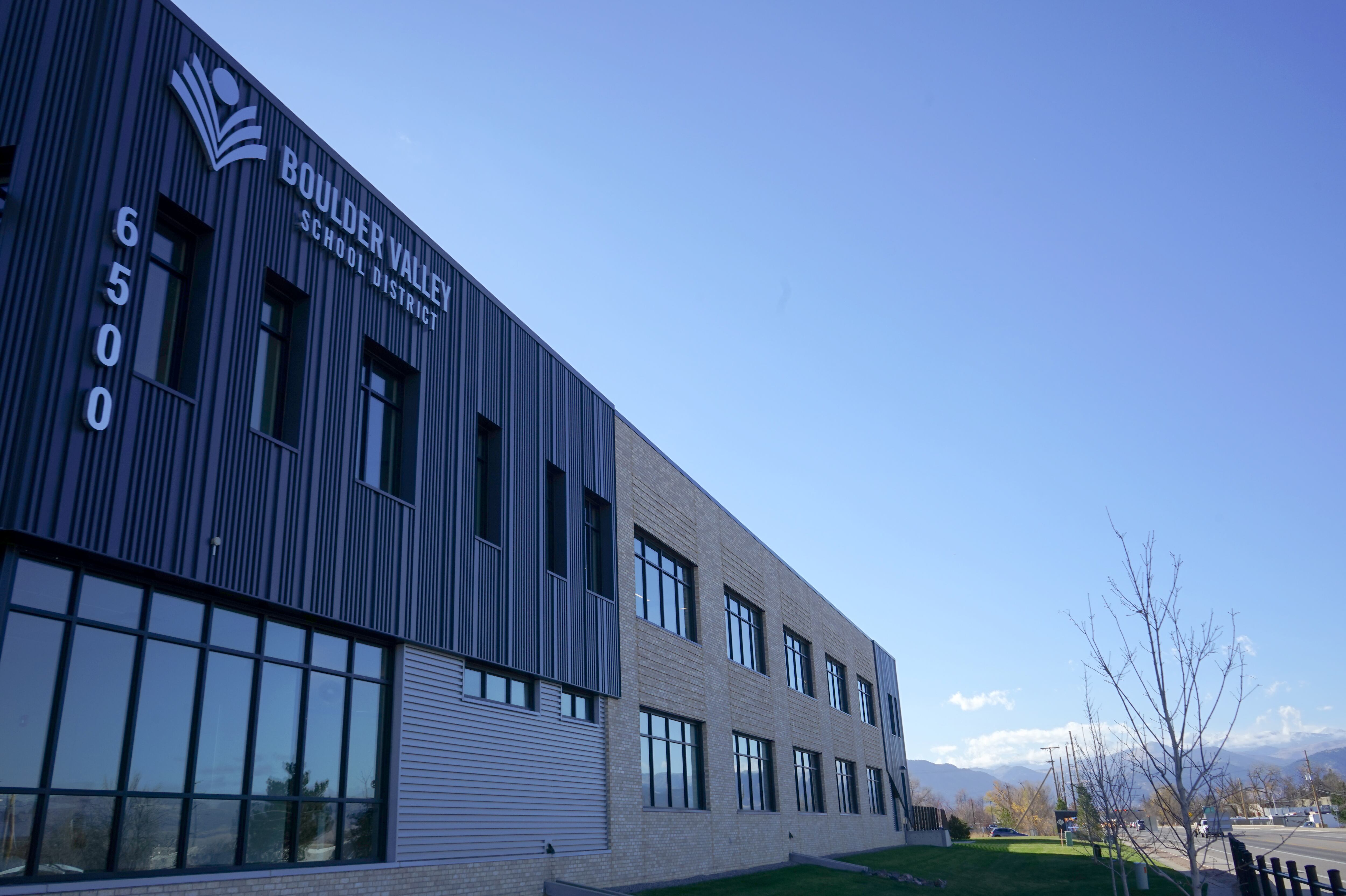 A large office building fills the left side of the frame with a sign reading “Boulder Valley School District.” Mountains, trees, and blue sky can be seen in the distance.
