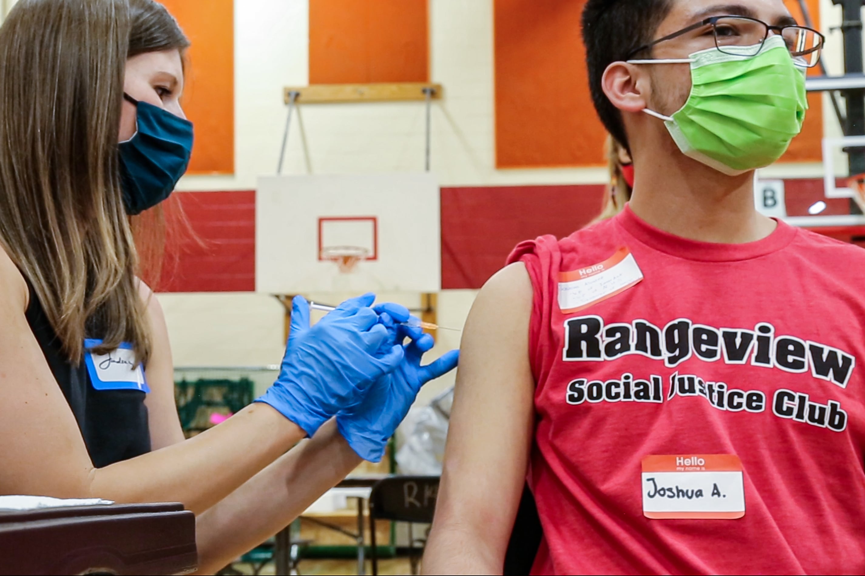 A woman in a mask administers a COVID vaccine to a teenage boy wearing a red Rangeview Social Justice Club T-shirt.
