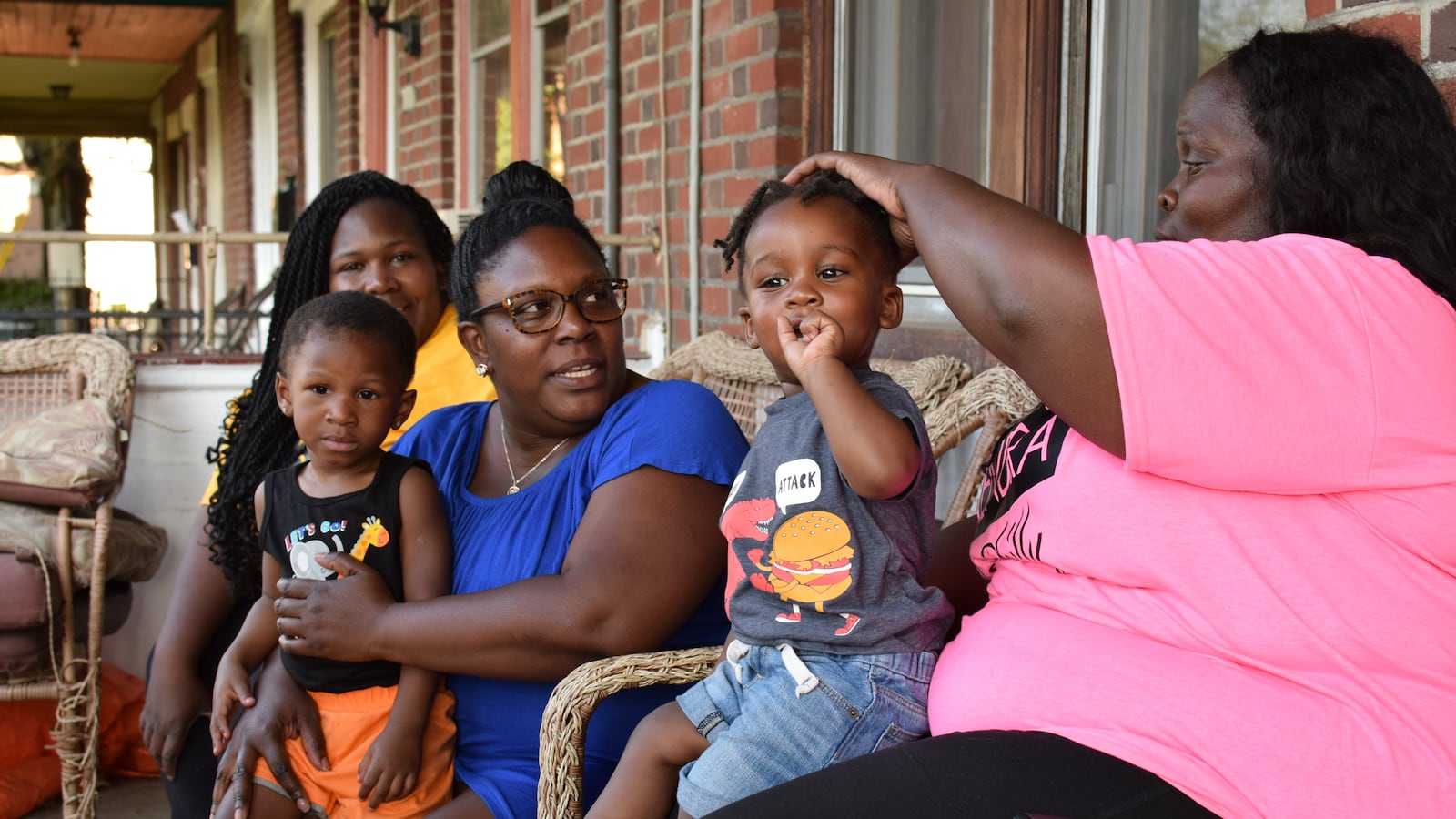 Three women sit on chairs outside of a red brick home with two young children sitting on their laps.