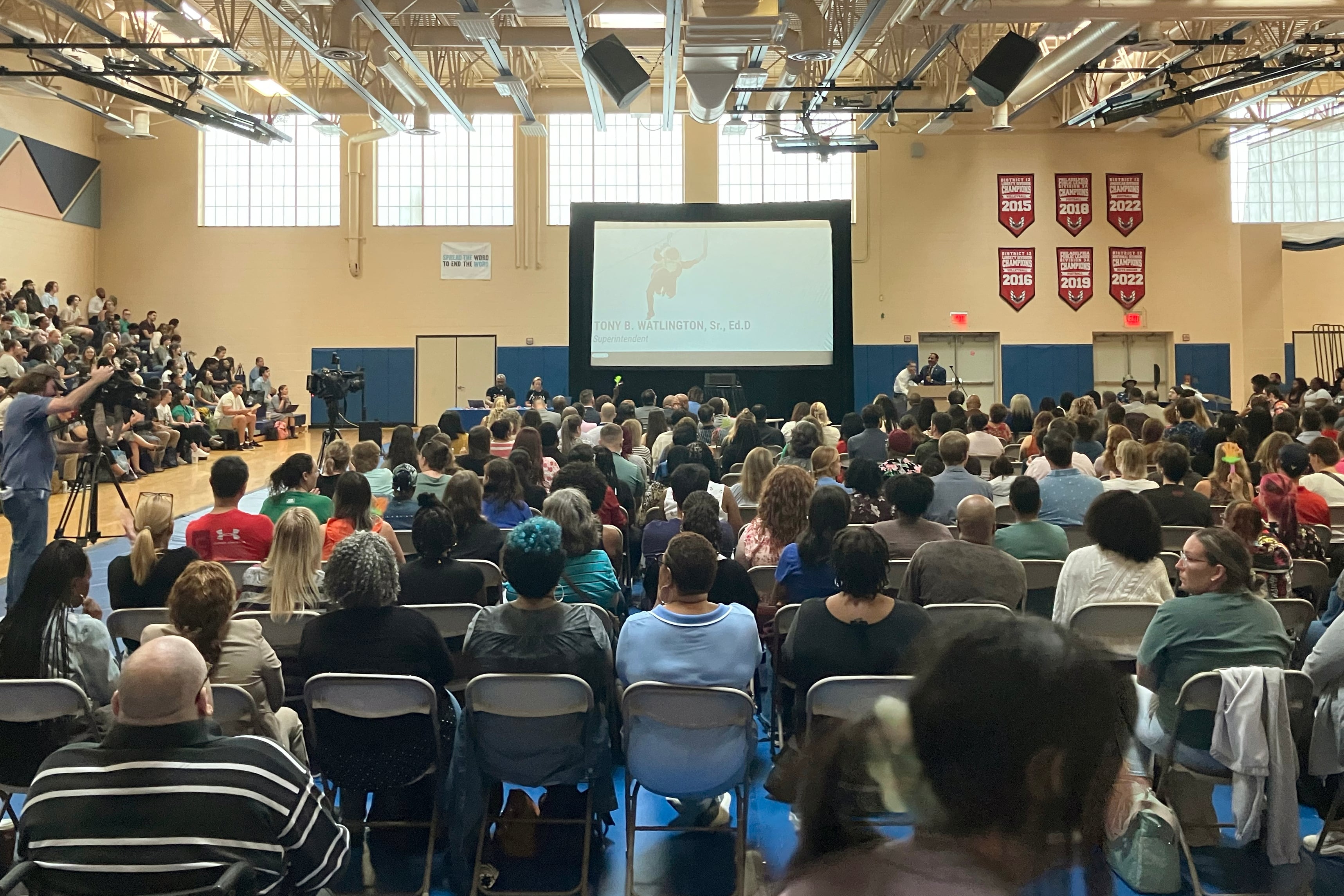 A room full of teachers, staff and others sit in a gymnasium with a large white projector screen in the background.