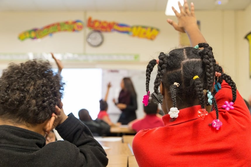 Two elementary students, shown from behind, sit in a classroom with a white teacher.