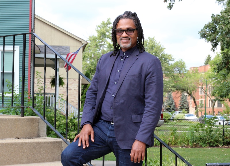A man with short dark braids and wearing a dark suit jacket poses for a portrait near a fence outside.