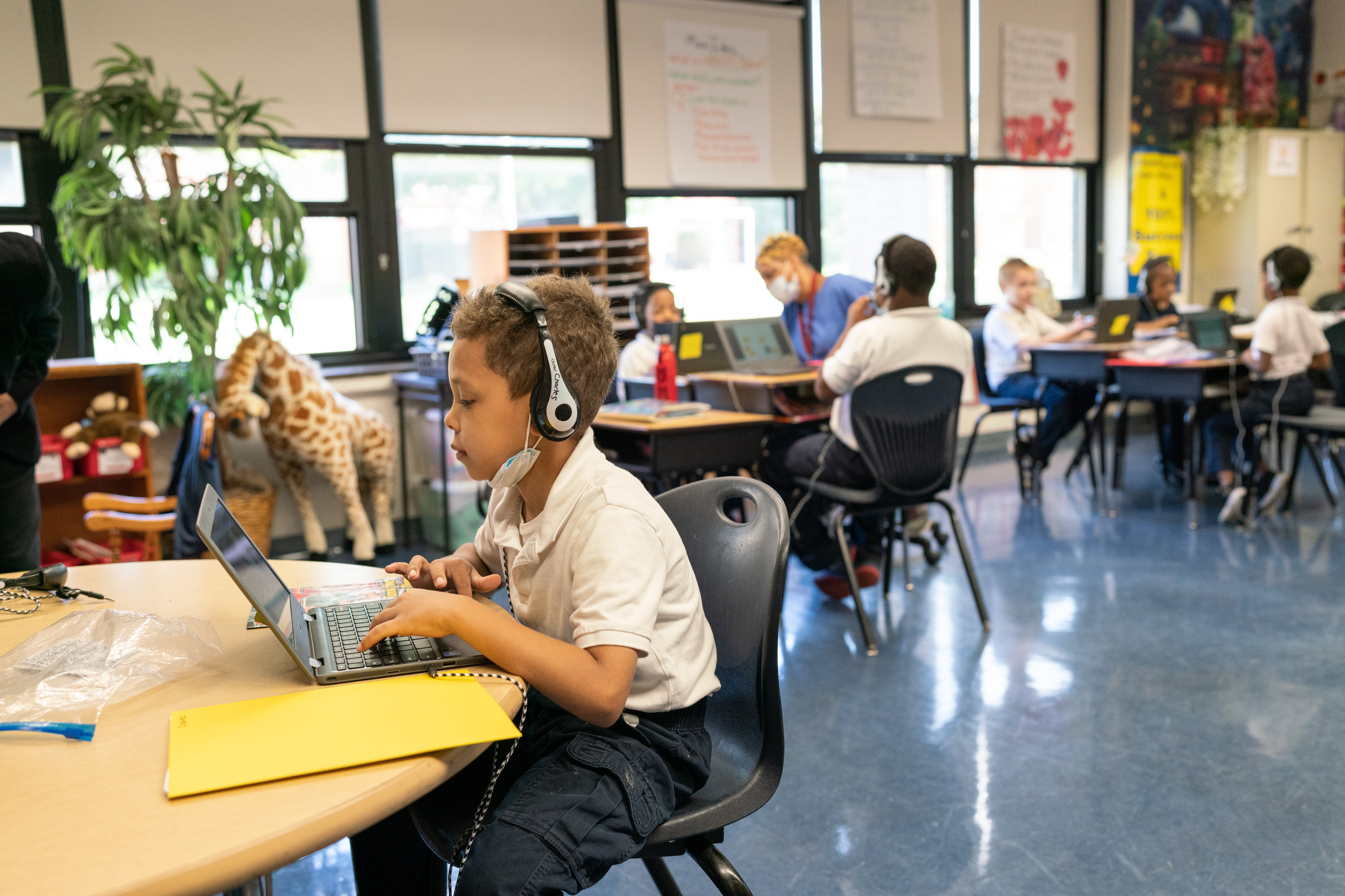 A student wearing headphones sits at a table in a classroom and works on a laptop. In the background, other students are sitting at desks.