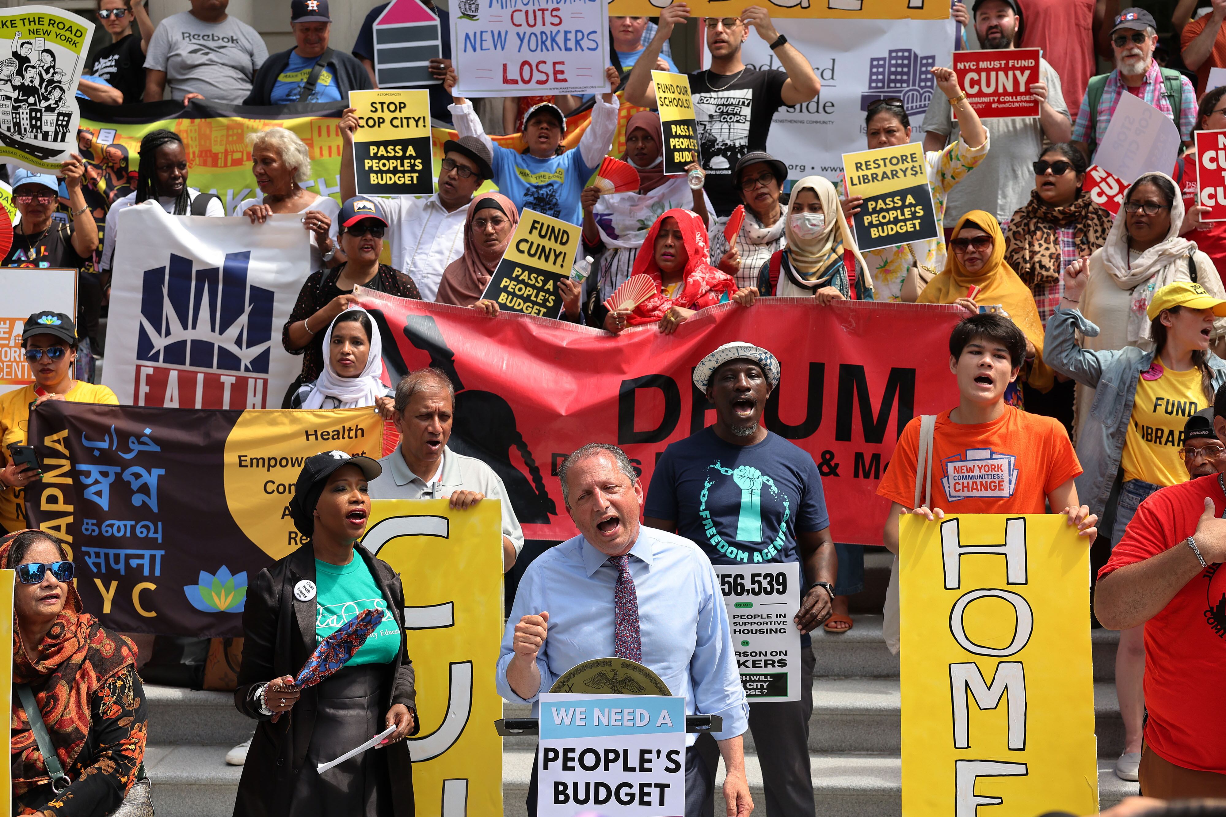 A man wearing a dress shirt and a tie speaks from behind a poster and in front of protestors during a rally at NYC City Hall.