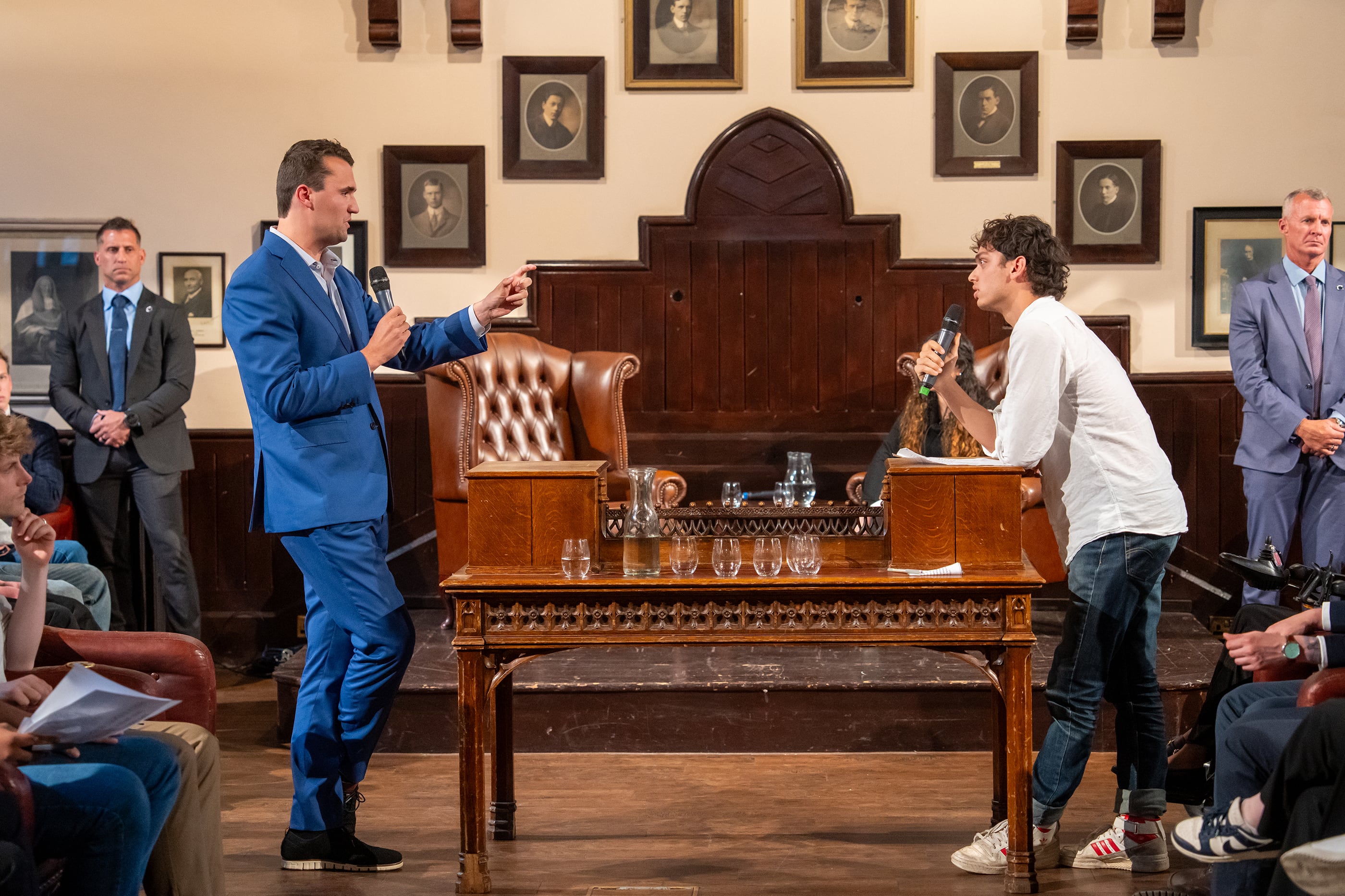 A photograph of a white man in a blue suit standing across a table from a college student wearing a long sleeve shirt both standing in a university room.