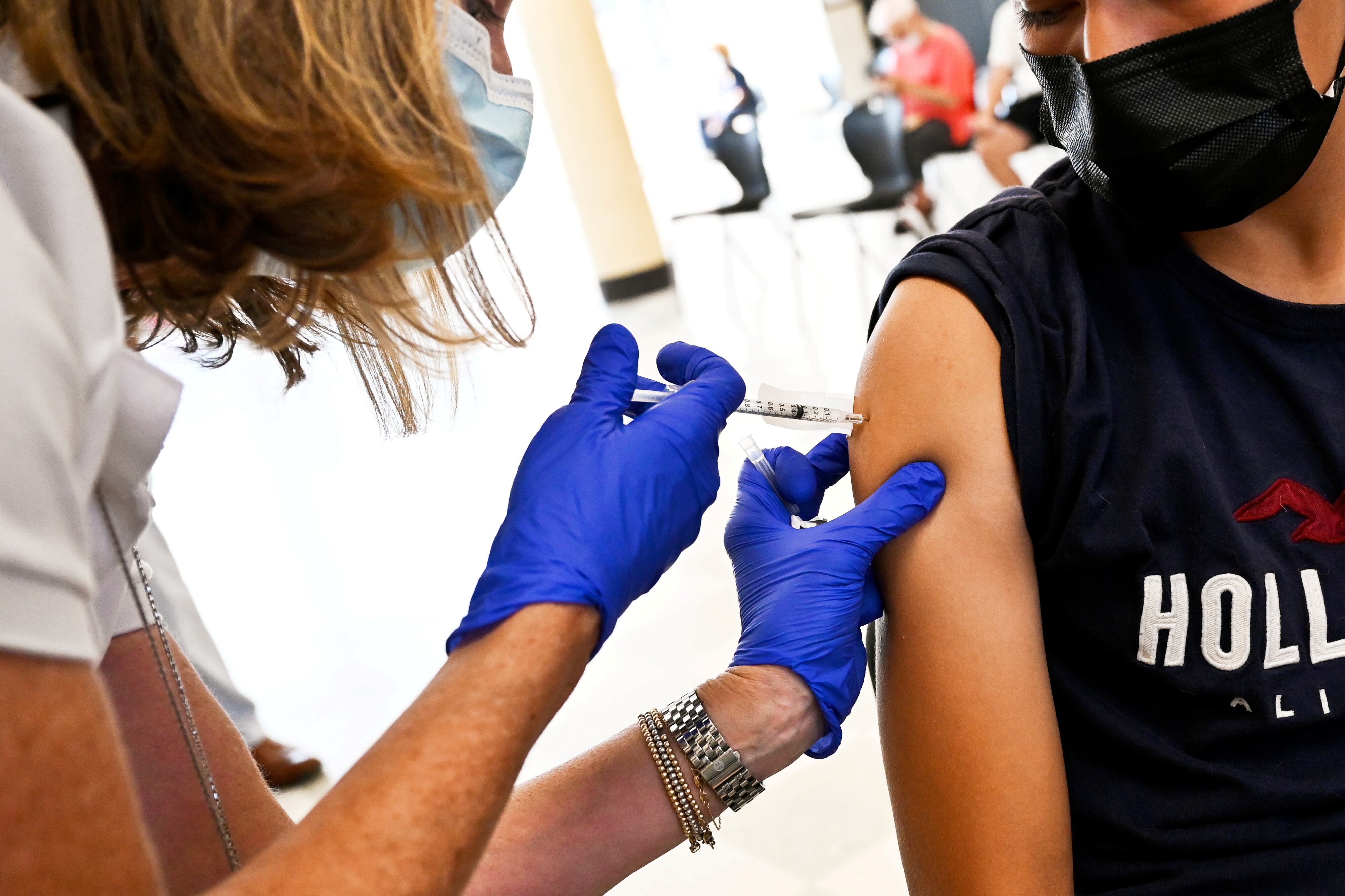 A student wearing a Hollister shirt receives a dose of a COVID vaccine from a health care professional wearing purple latex gloves.