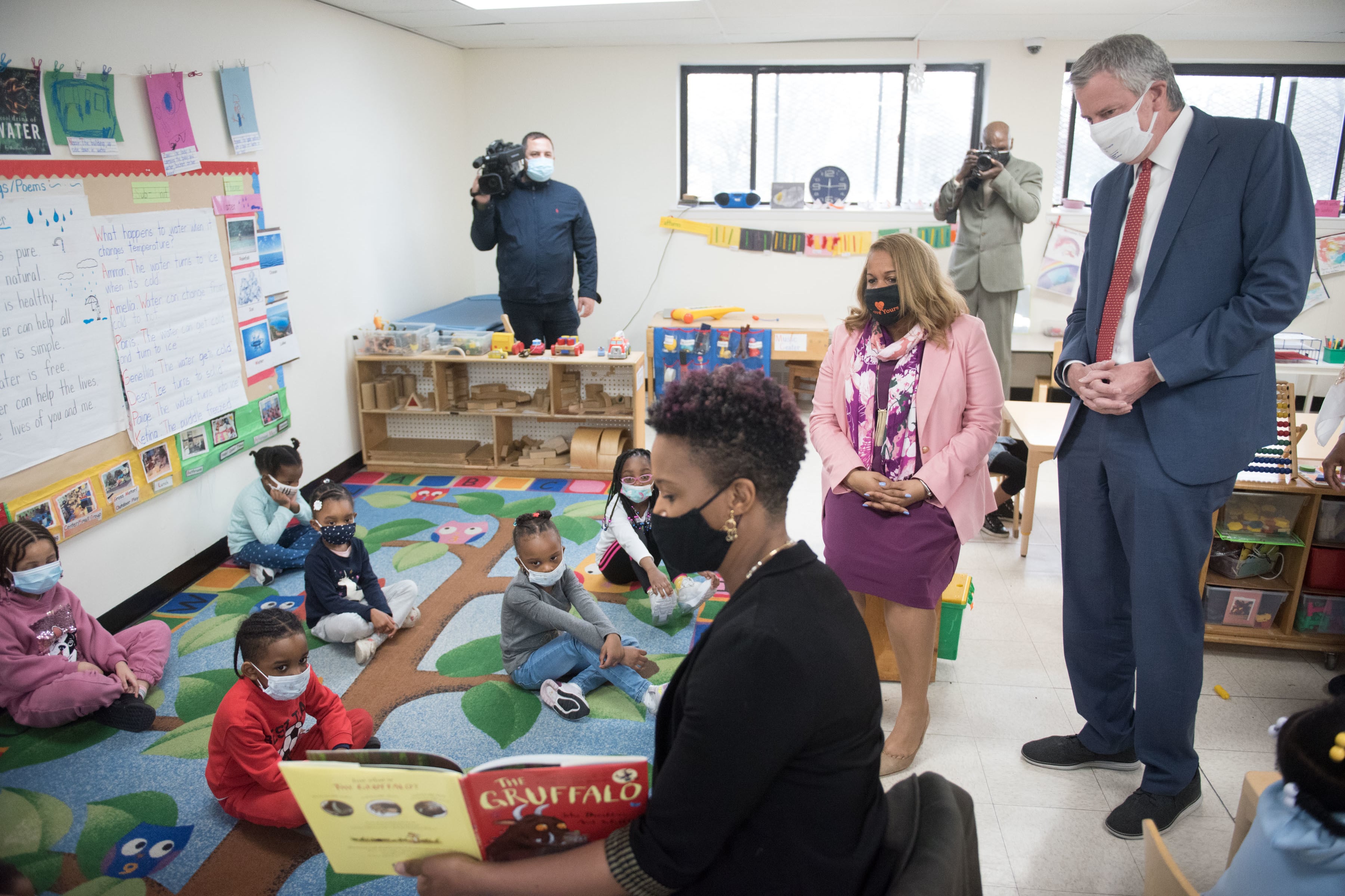 Mayor Bill de Blasio and Chancellor Porter look on as a teacher reads to a group of five students on a colorful rug. The teacher with the book is in the foreground, while the mayor and chancellor are to the right.