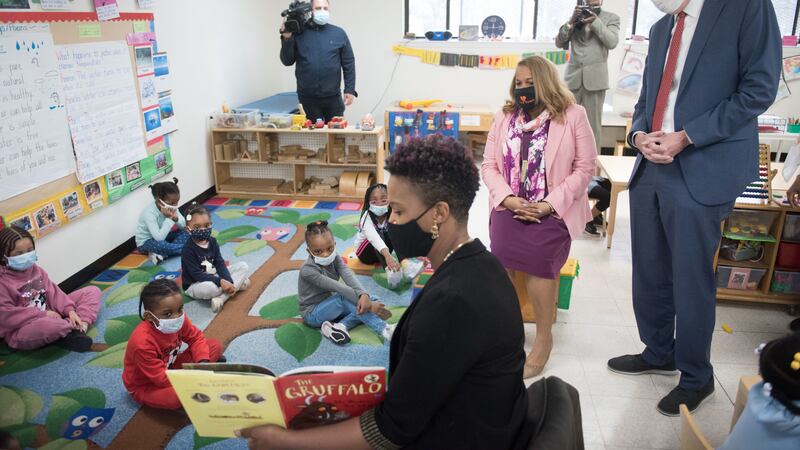 Mayor Bill de Blasio and Chancellor Porter look on as a teacher reads to a group of five students on a colorful rug. The teacher with the book is in the foreground, while the mayor and chancellor are to the right.