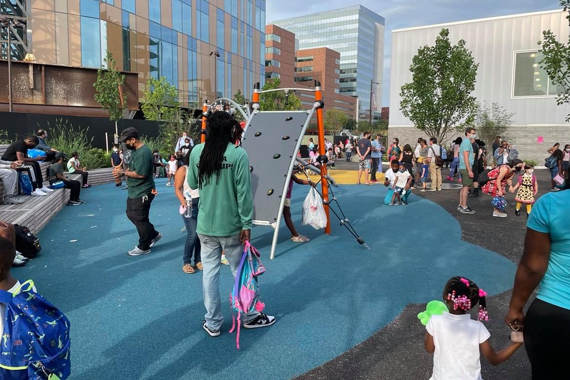 A parent in a green shirt and jeans holds a backpack in the middle of a play yard at a new school. Other parents and children can be seen in the distance.