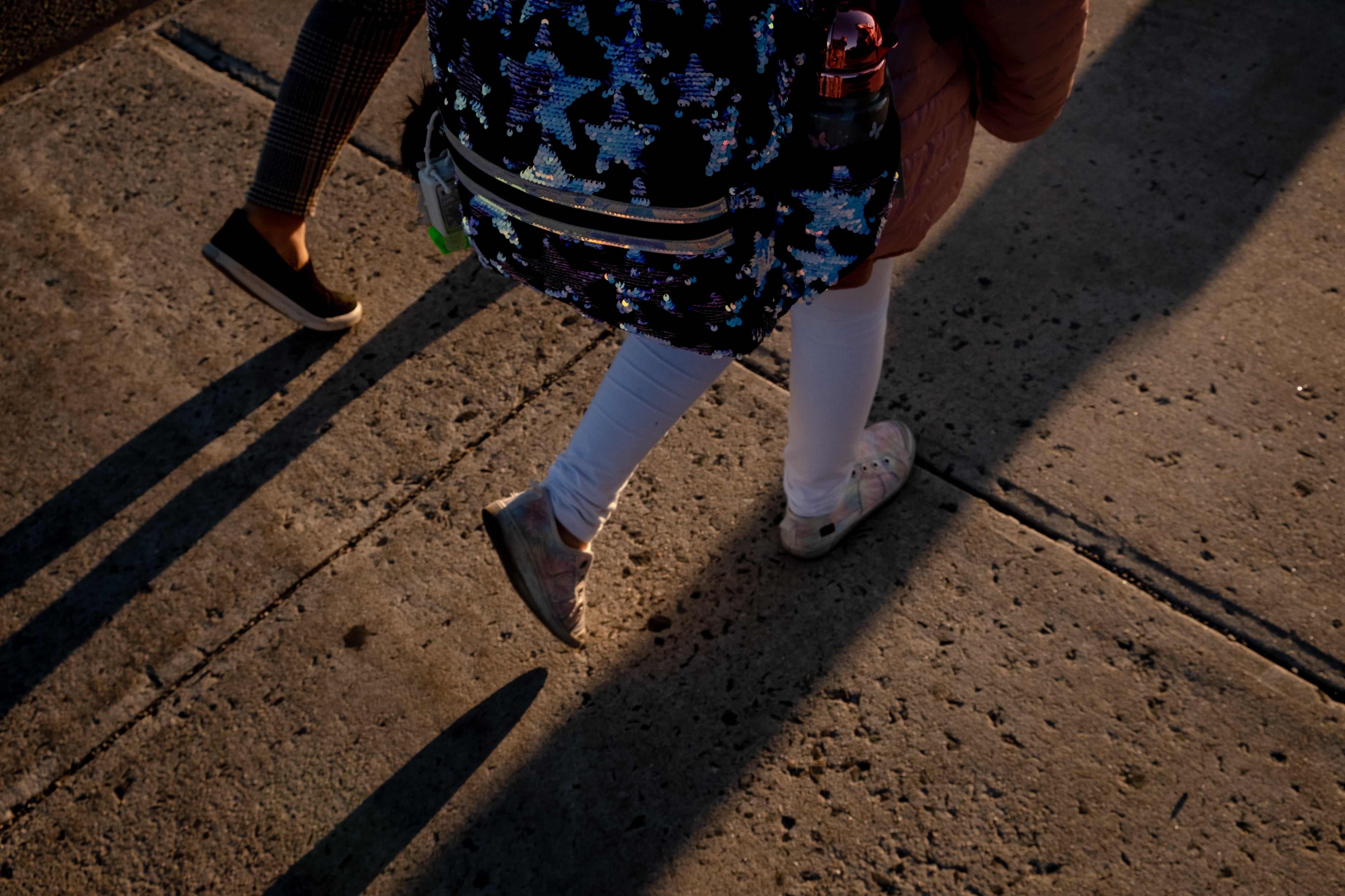 A view of a young student's legs and backpack while they hold the hand of an adult walking down the street.