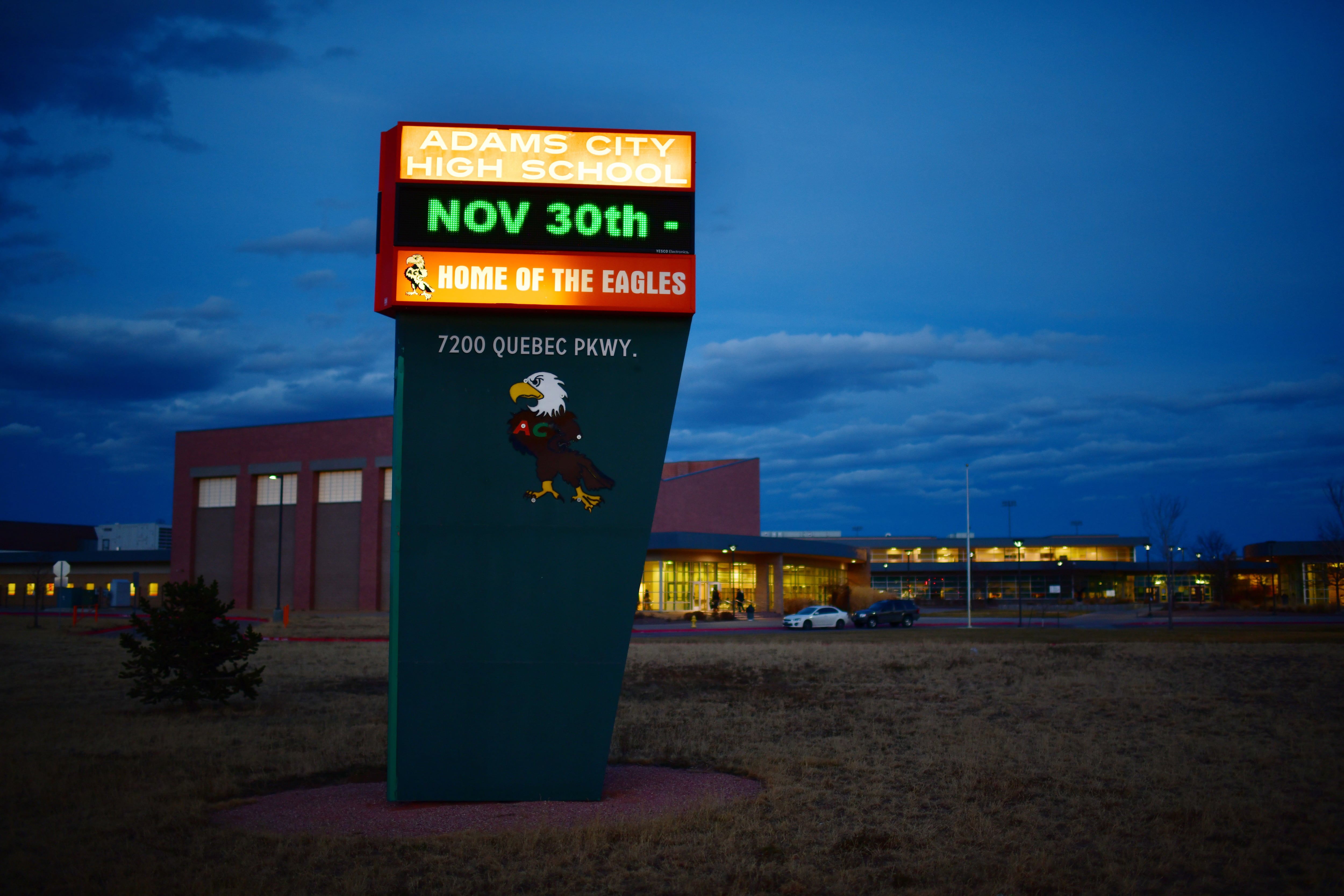 A brightly lit sign sits in front of Adams City High School against a dark blue sky.