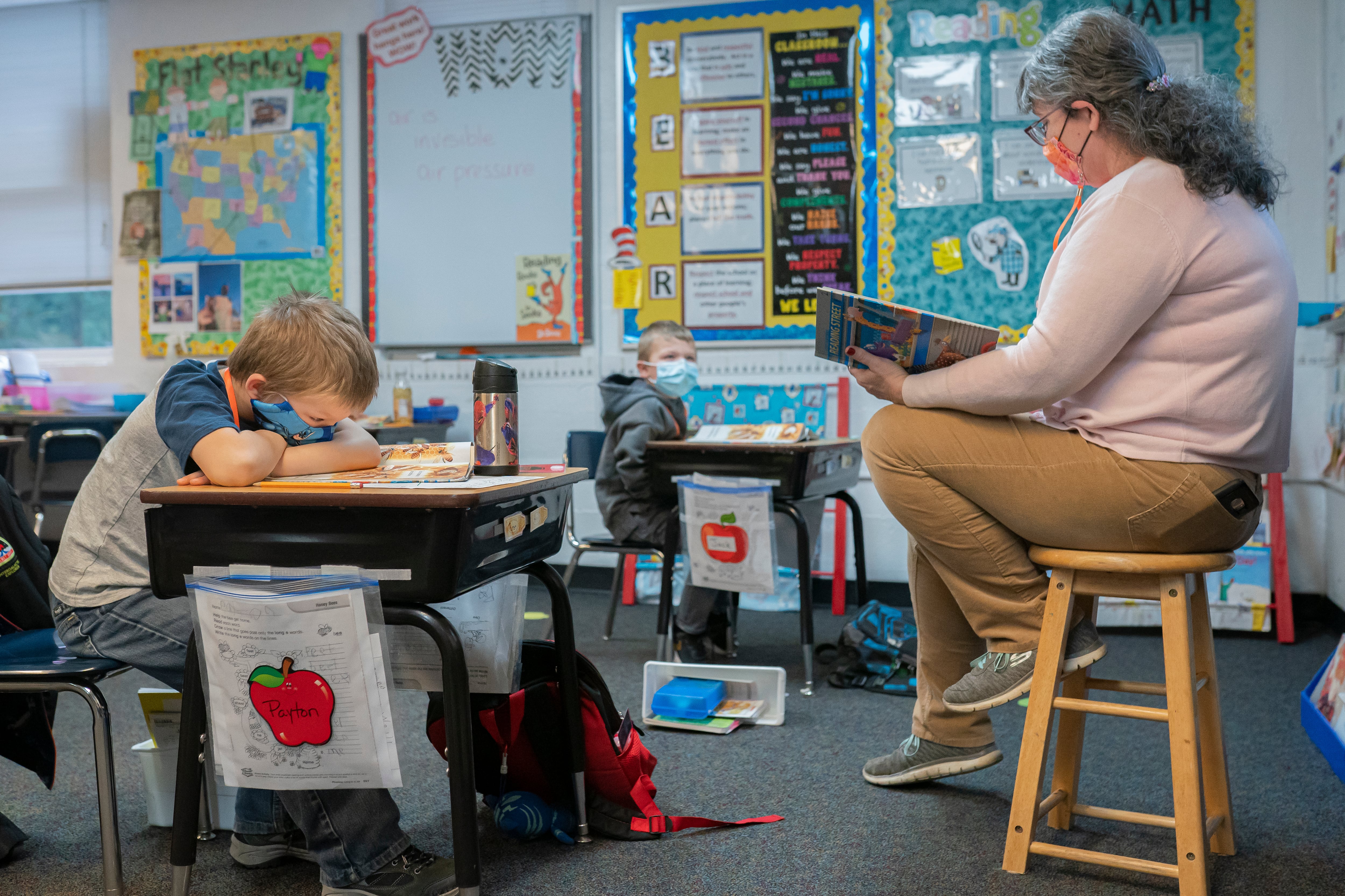 A masked teacher sitting on a stool looks at a book while a masked student, sitting at a desk in front of the teacher and his head resting on his crossed arms, looks at an open book. 