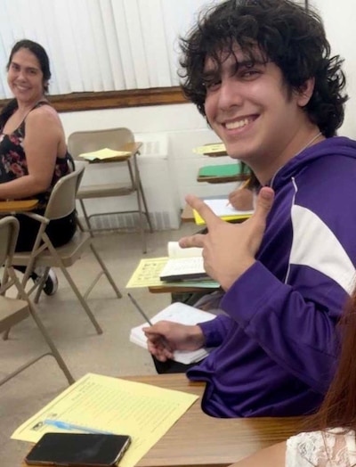 A photograph of a high school student sitting at a desk posing for the camera while a woman in the back sits at a desk and smiles at the camera.