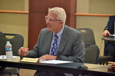 A man with white hair and wearing a gray suit sits at a table in a conference room.