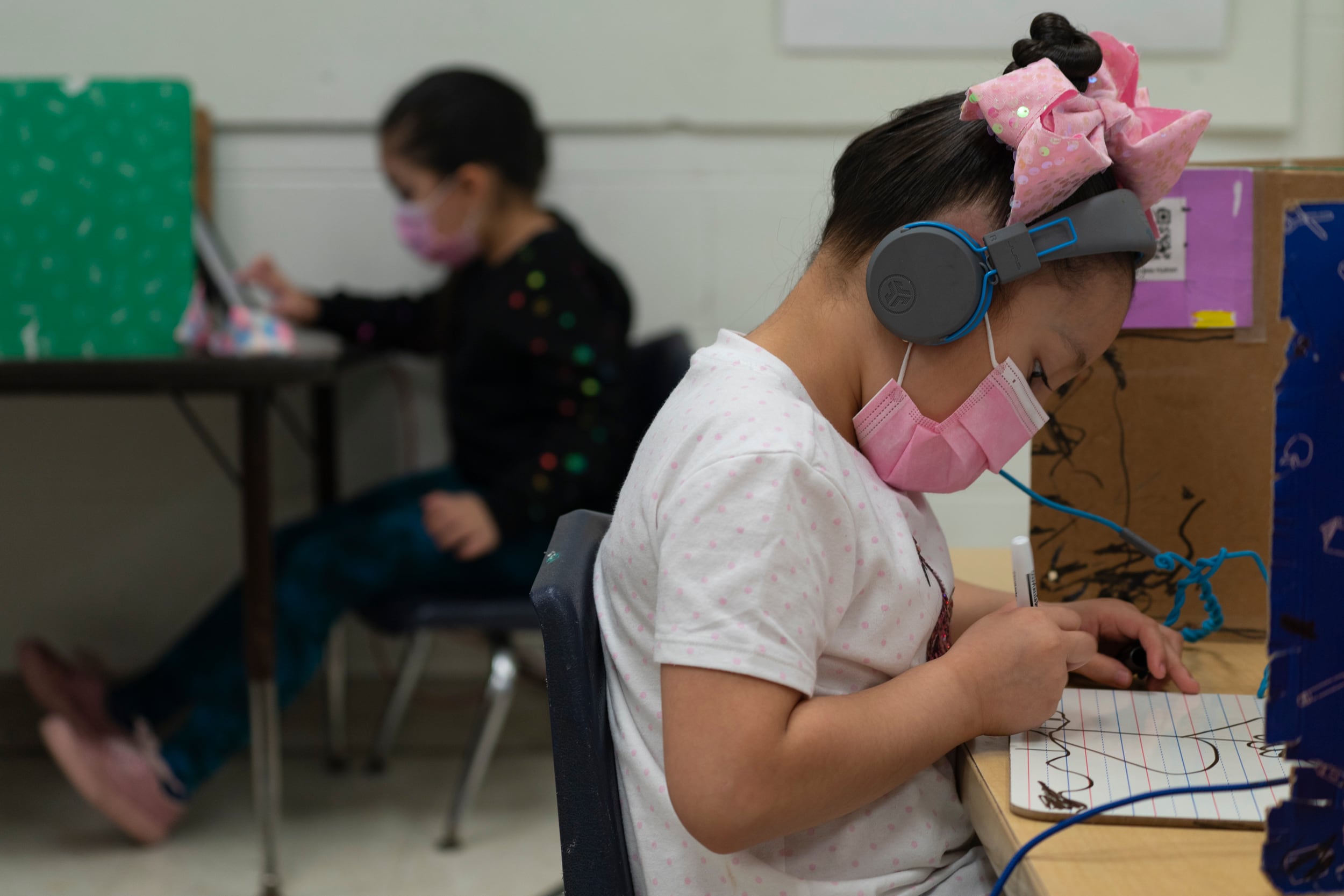 A student wearing a pink bow and protective mask draws on a whiteboard at her desk.