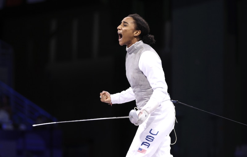 A young woman wearing a fencing uniform holds a fencing sword and screams with joy with a black background.