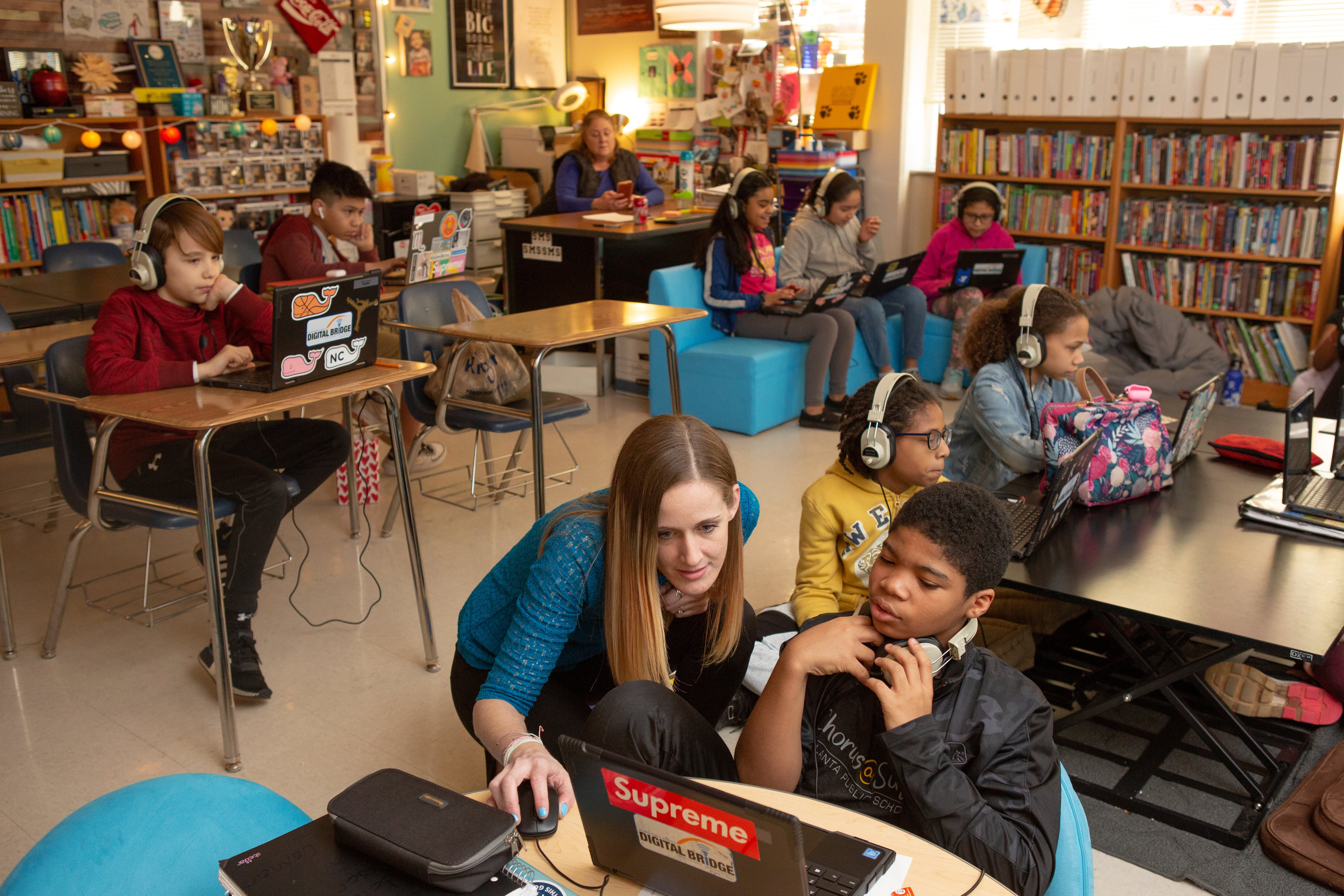 An adult wearing a blue blouse helps a student on a laptop in the foreground while other students sit at their desks working on laptops. Shelves with books line one wall of the classroom.
