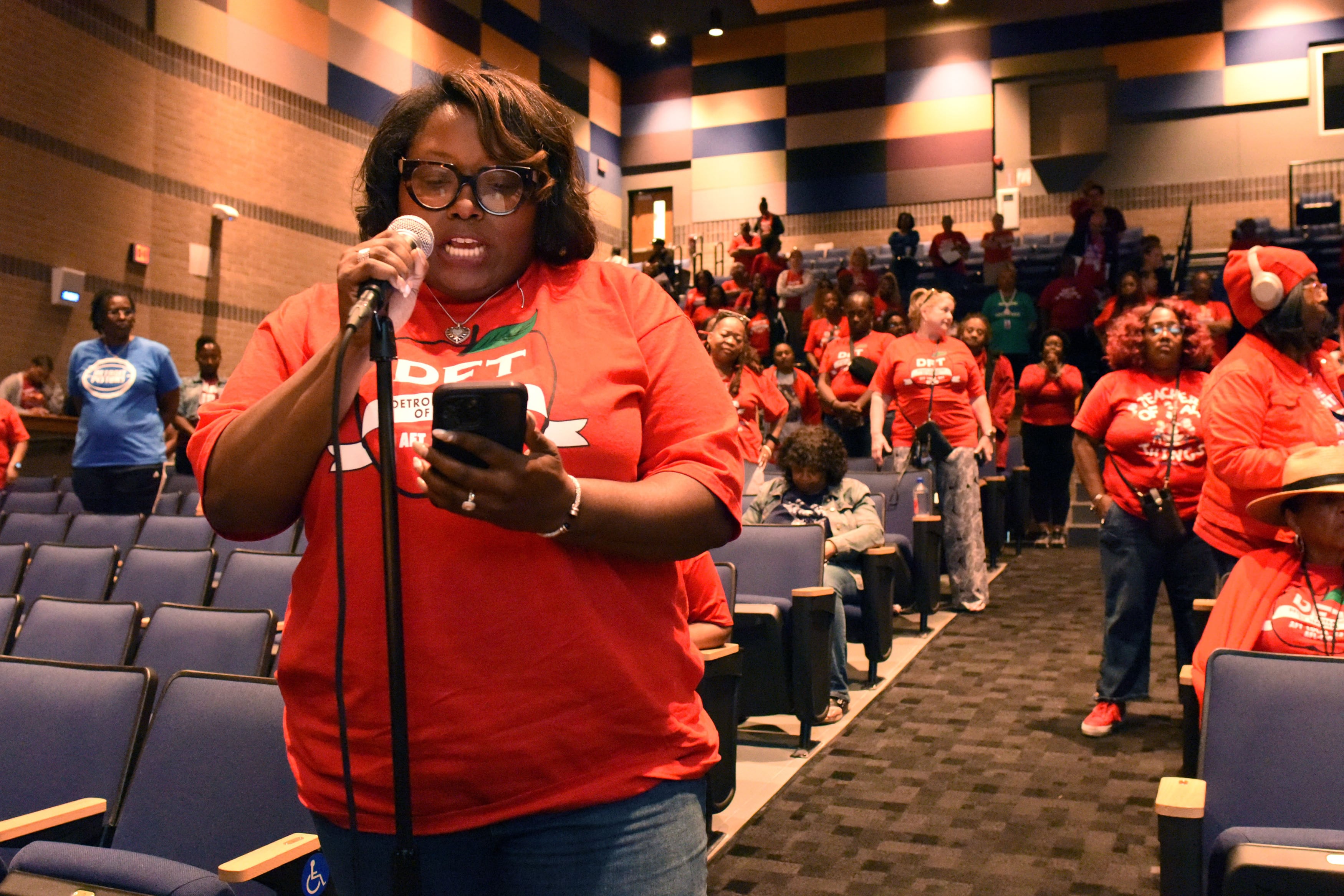 A woman with short dark hair and wearing a red t-shirt holds a cellphone while speaking into a microphone with a large group of people sitting and standing behind her.