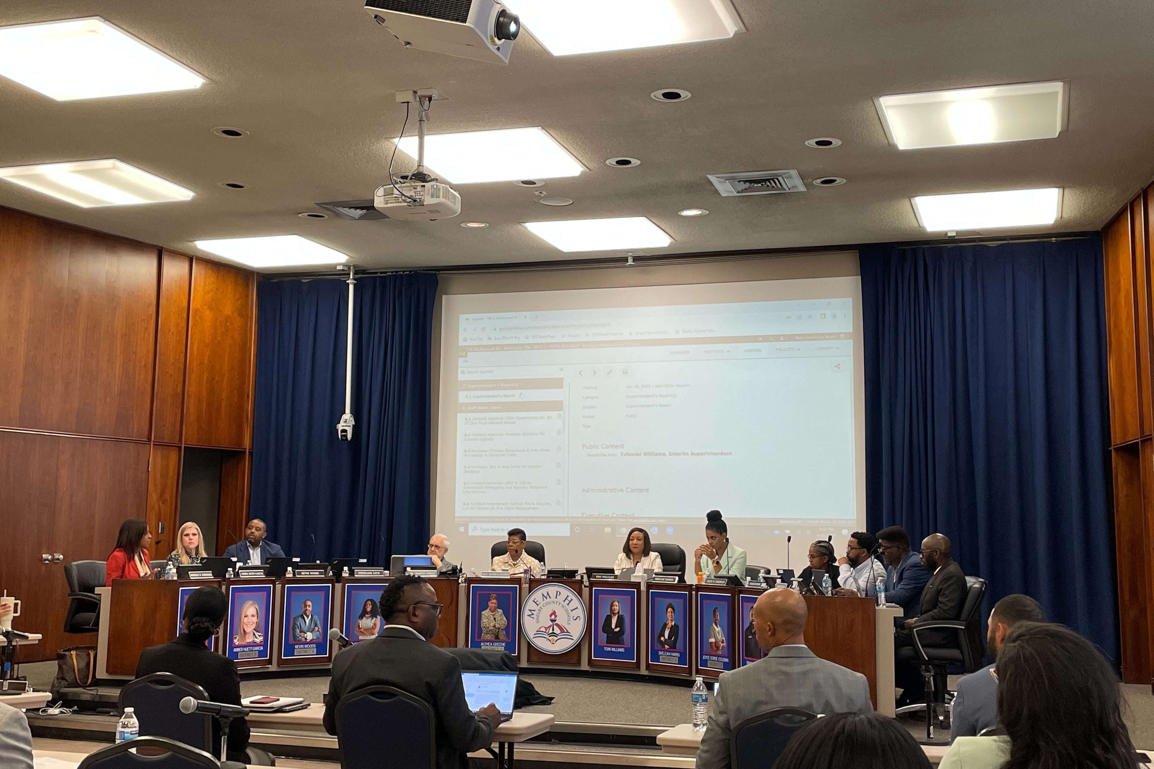 Eleven people sit around the dais in front of a projector surrounded on either side by navy curtains.