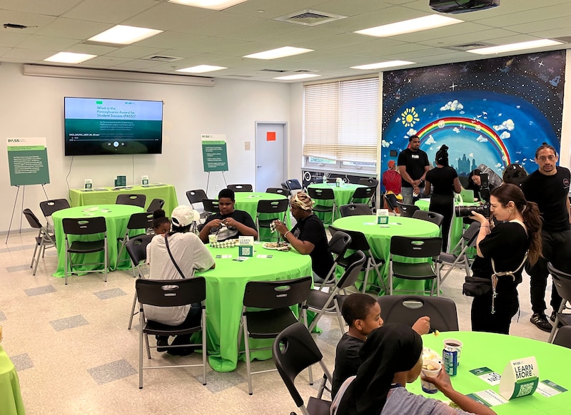 A group of children sit at different tables with green table cloths.