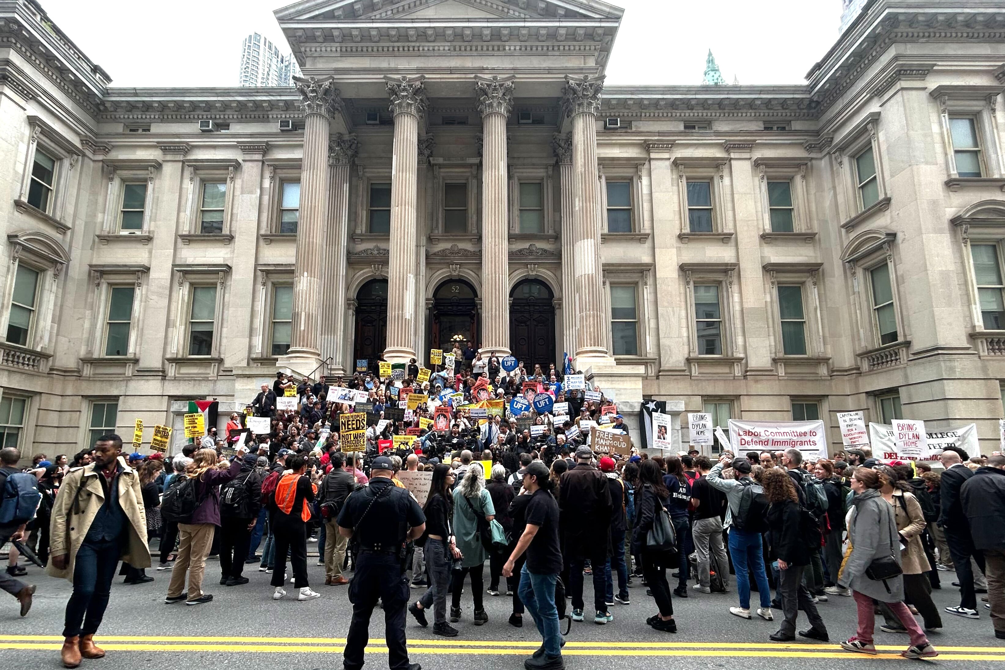 A crowd of people, some with signs, stands on the steps of a building.