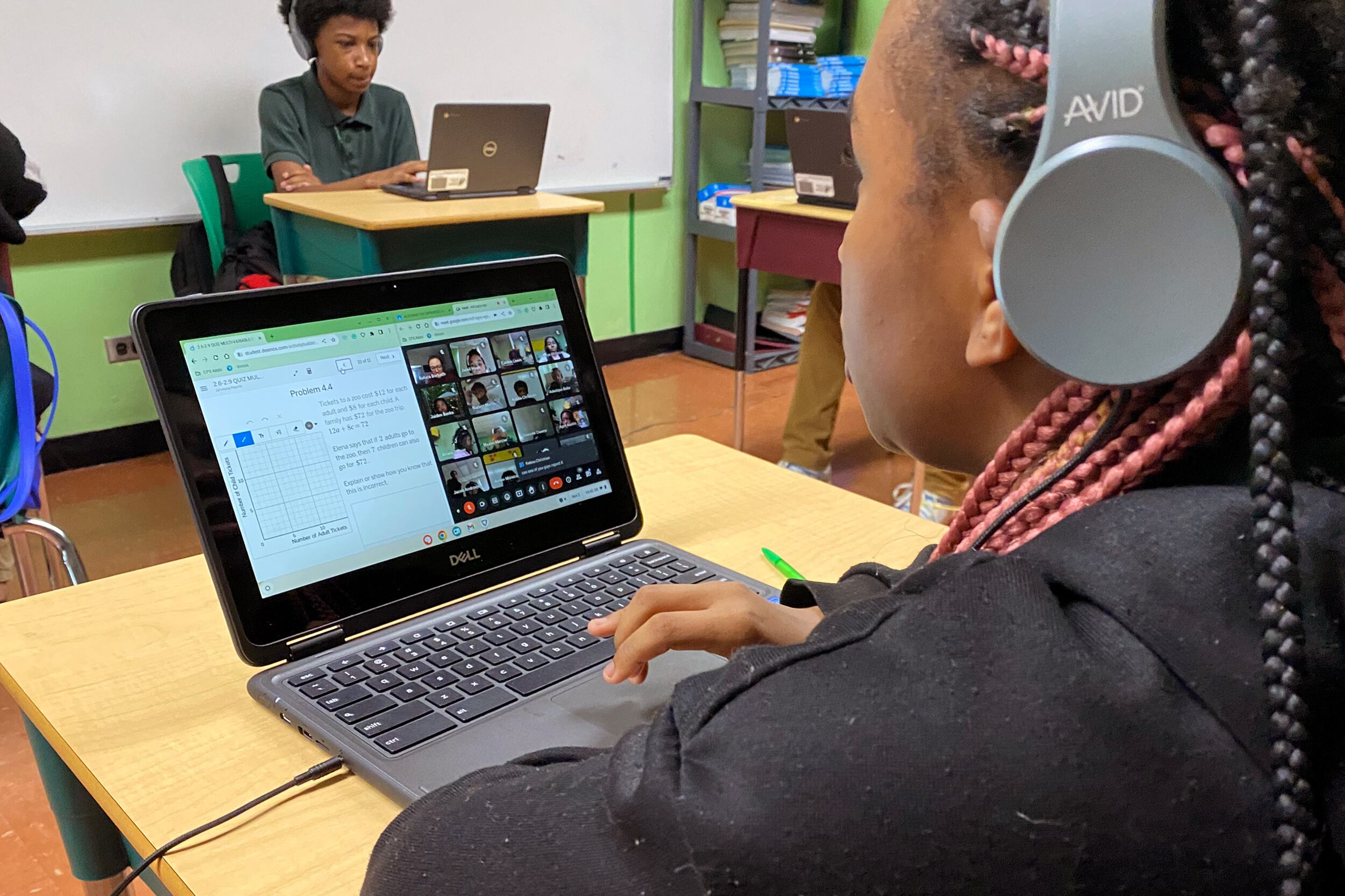 A student with long black and pink braids works on a computer at her desk while a student works on a computer in the background in a classroom.