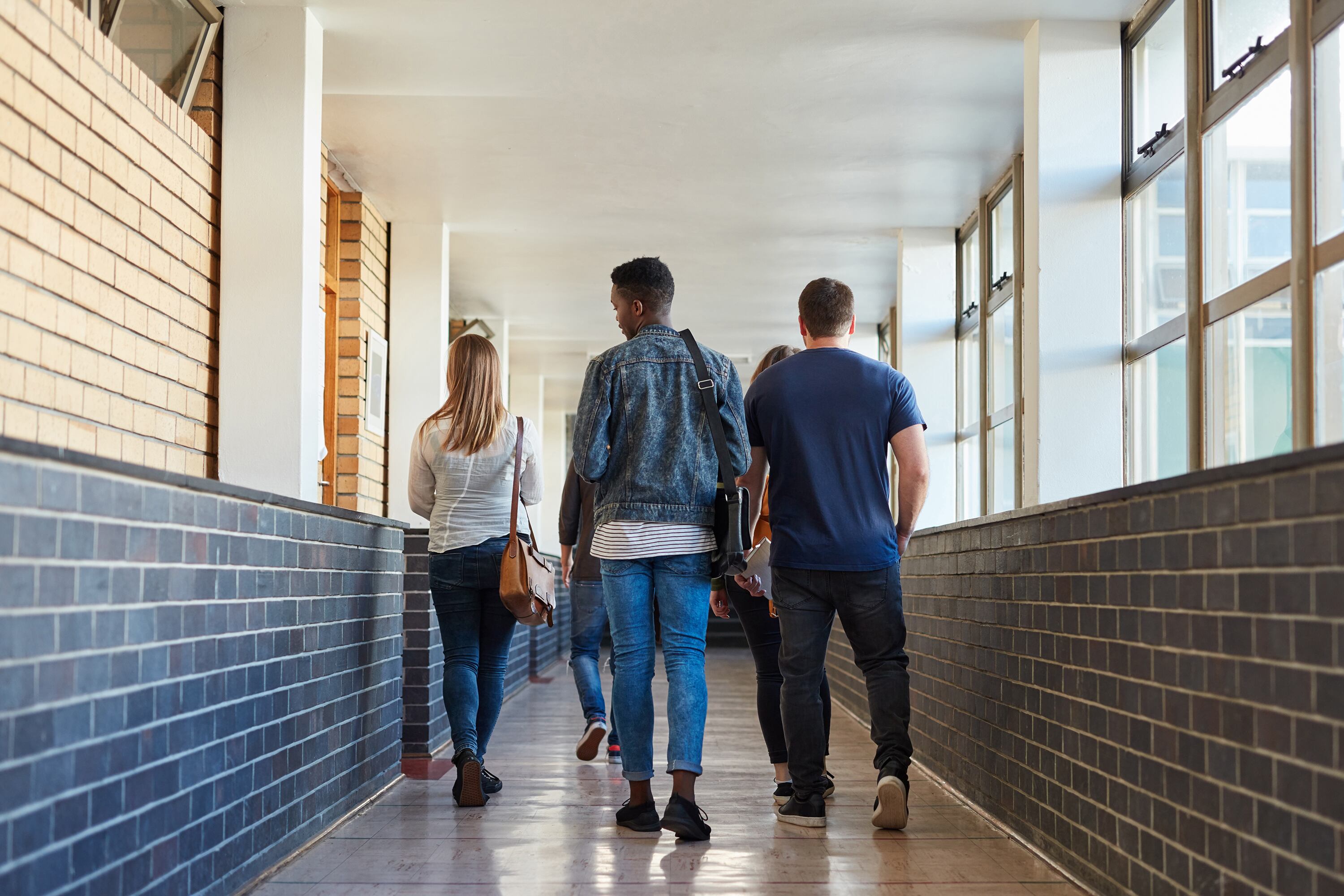 A photo from behind four high school students walking down a well lit hallway with a brick wall on the left and windows on the right.