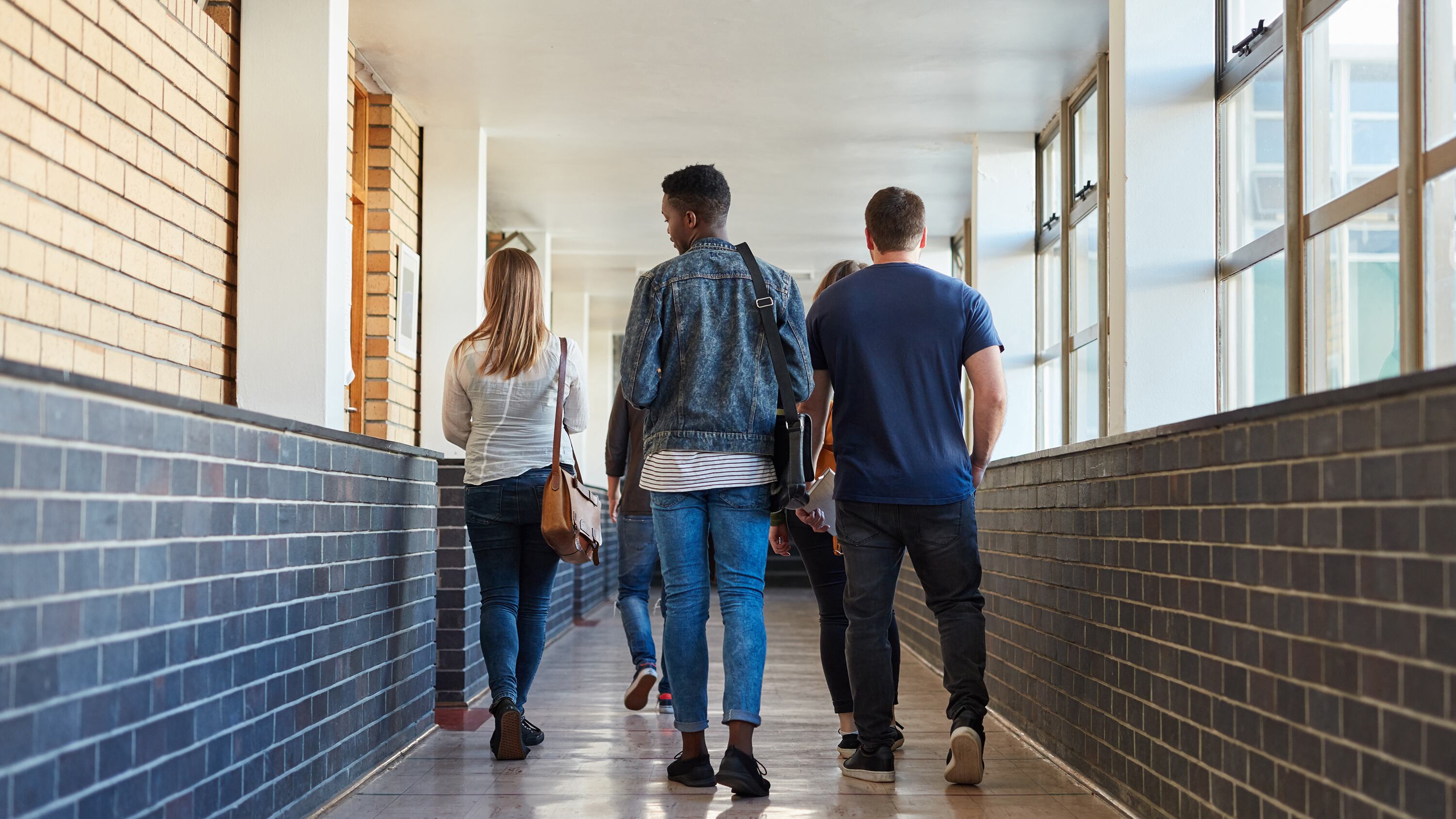A photo from behind four high school students walking down a well lit hallway with a brick wall on the left and windows on the right.