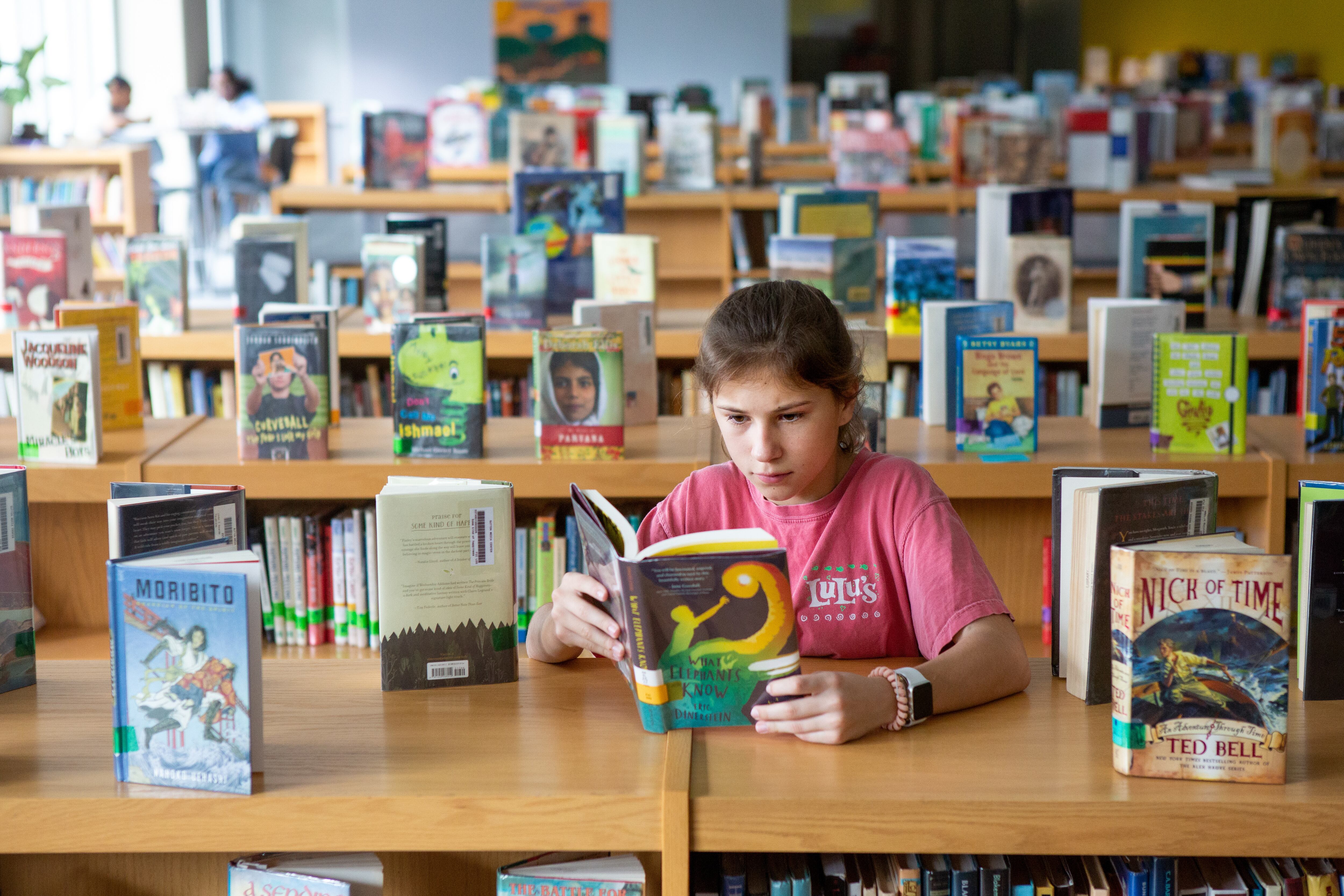 A child sits at a table reading a book in a room filled with tables covered in books. 