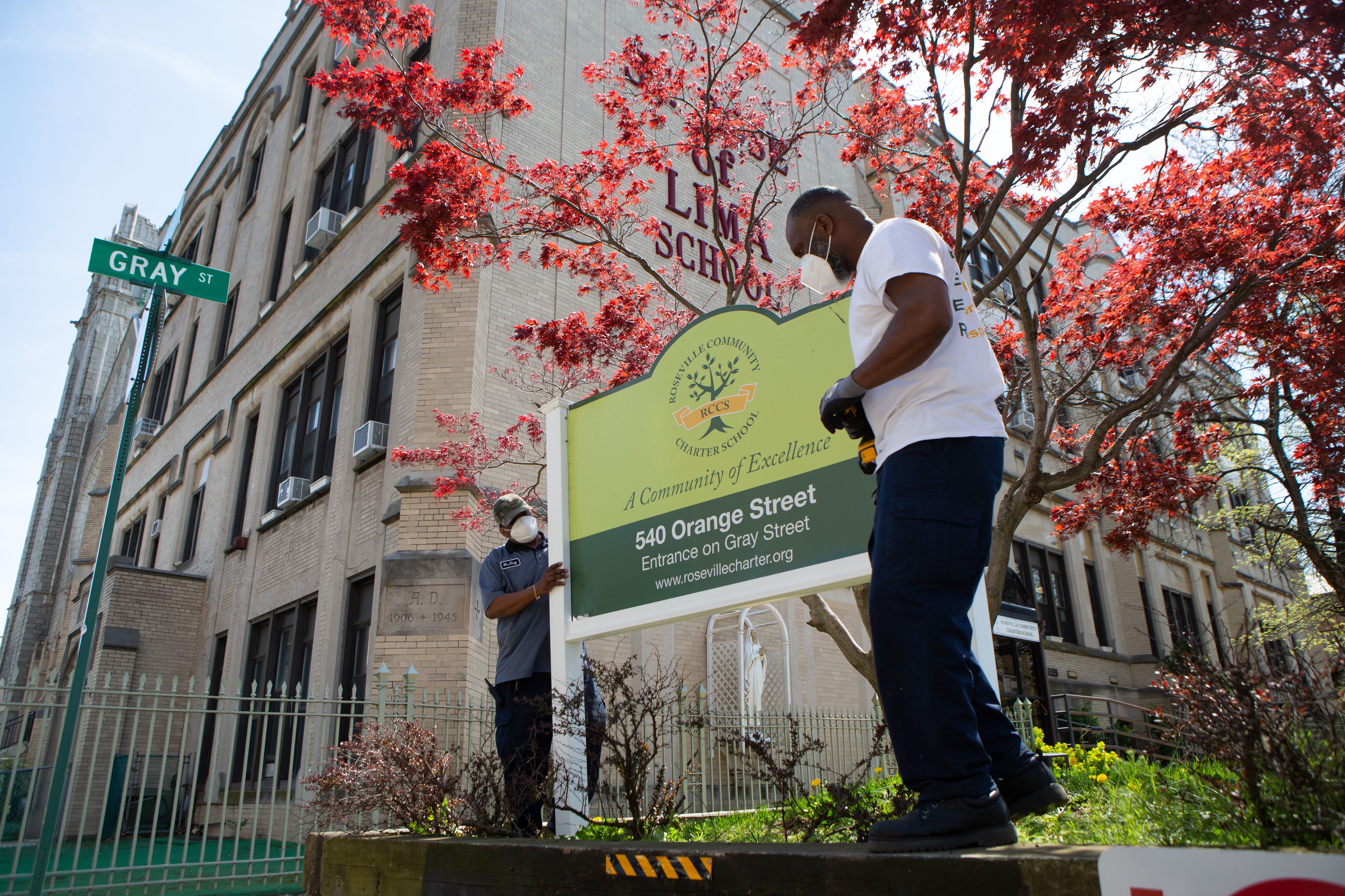 Two men wearing protective masks fix a sign on a bright Spring day outside of Roseville Community Charter School in Newark, N.J.
