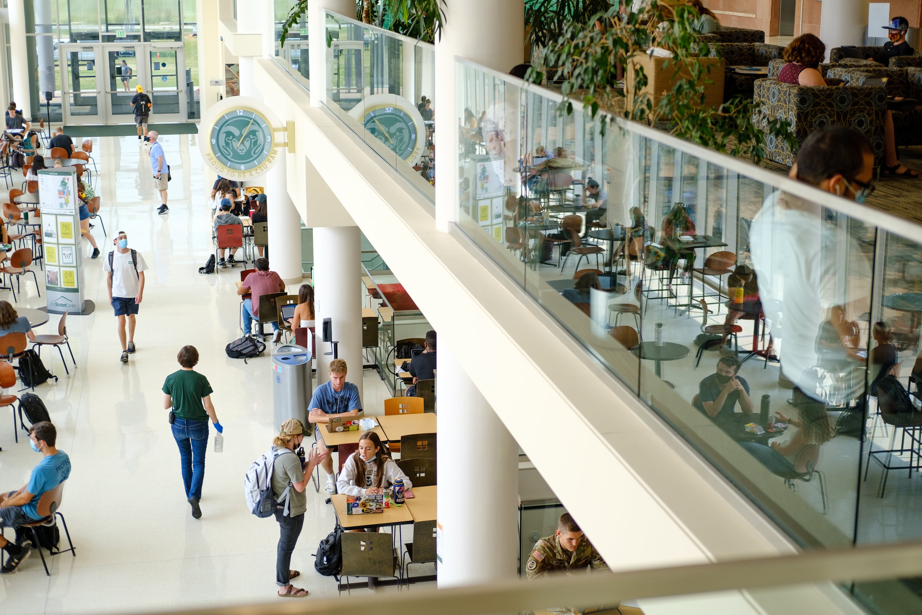 Students congregate in a student center, tables reflected off of a pane of glass in the building’s second level. A Colorado State University logo hangs above the columns connecting the two levels.