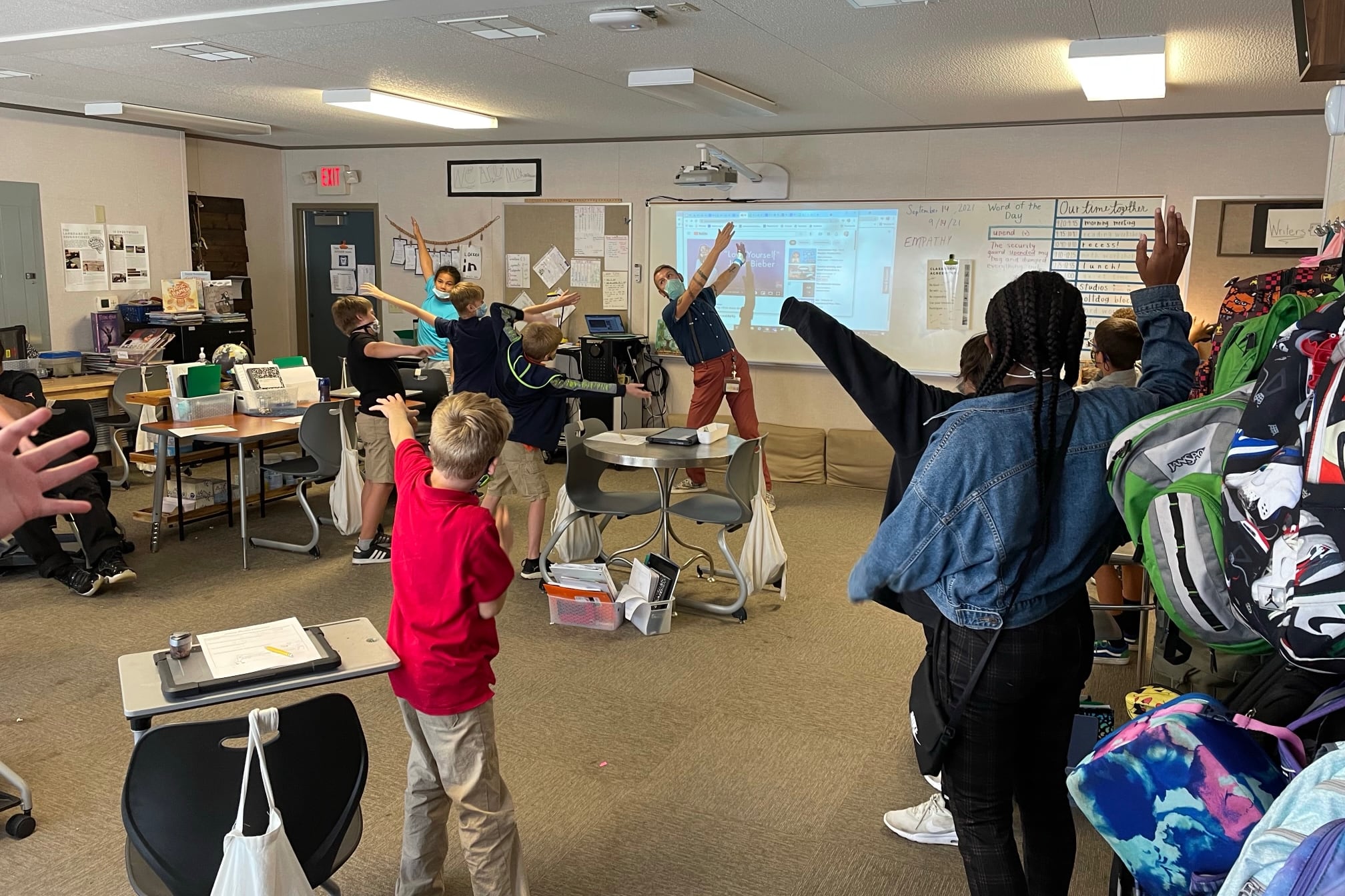 A teacher wearing a mask stands at the front of the classroom, swinging his arms in the air to demonstrate a calming strategy. Students scattered throughout the classroom wave their arms, too.
