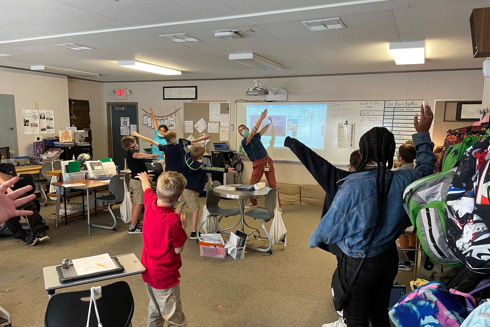 A teacher wearing a mask stands at the front of the classroom, swinging his arms in the air to demonstrate a calming strategy. Students scattered throughout the classroom wave their arms, too.