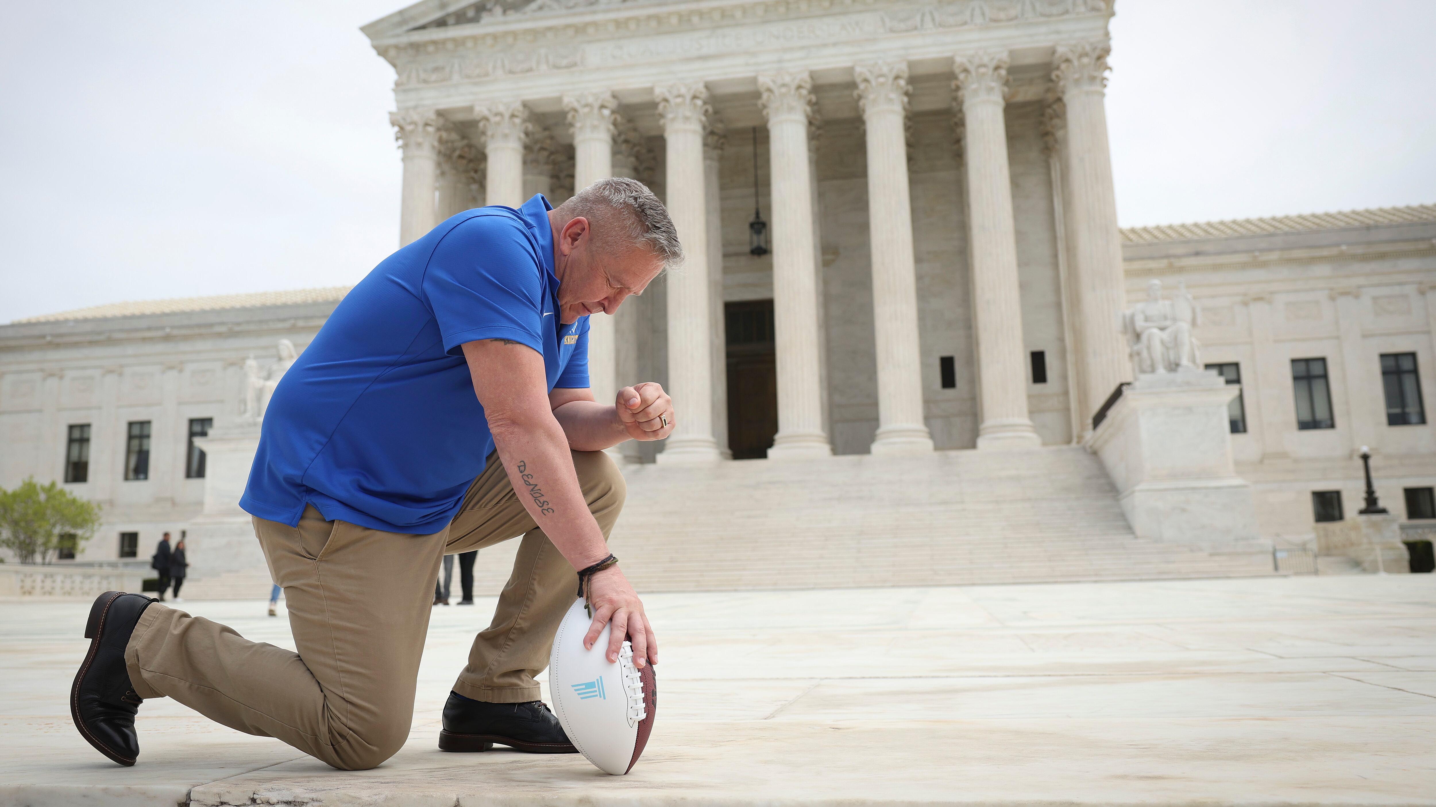 A man in a blue shirt prays in front of the United States Supreme Court building, holding a football.