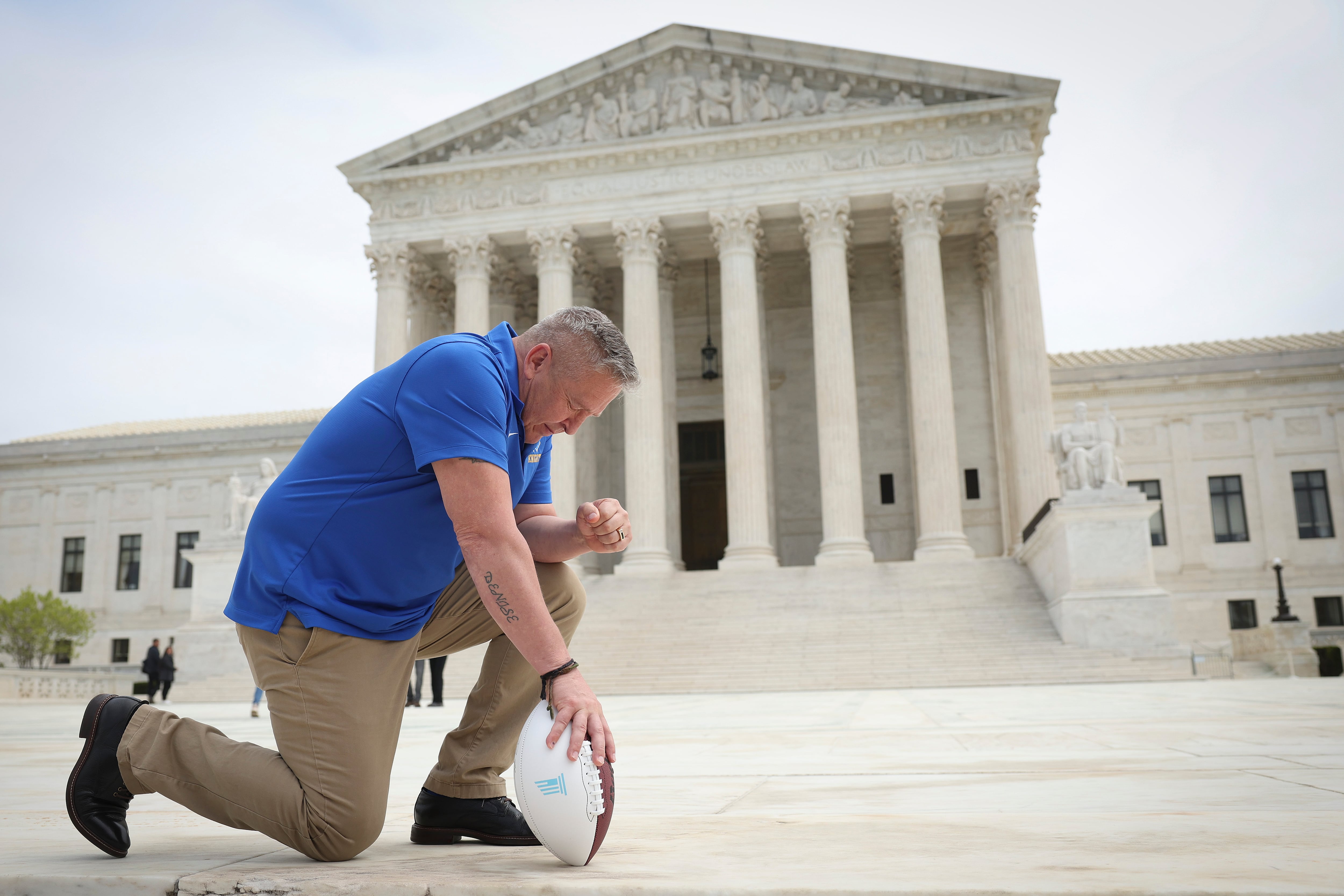 A man in a blue shirt prays in front of the United States Supreme Court building, holding a football.