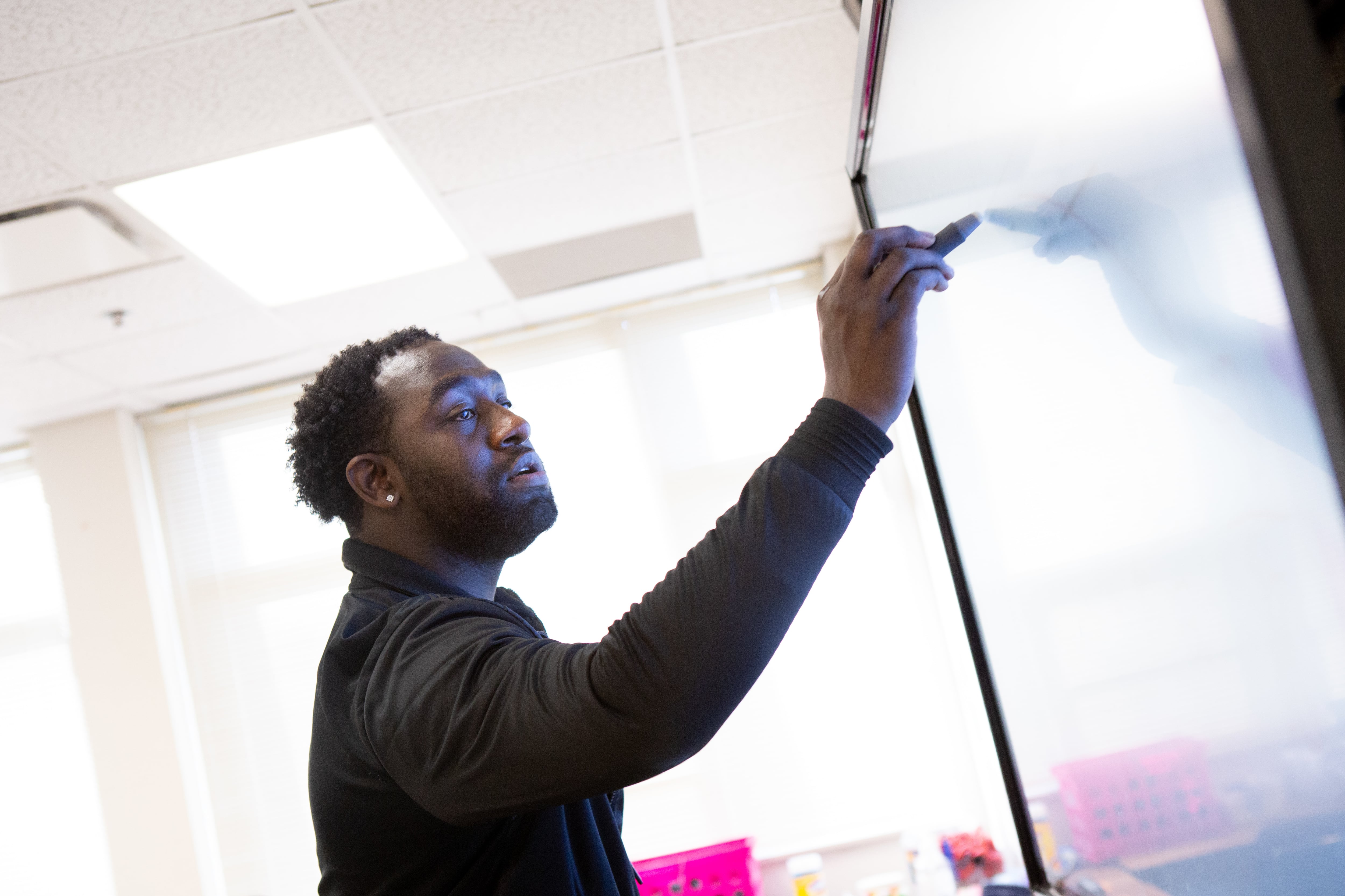 A teacher writes on a white board with a marker inside a bright sunny classroom.