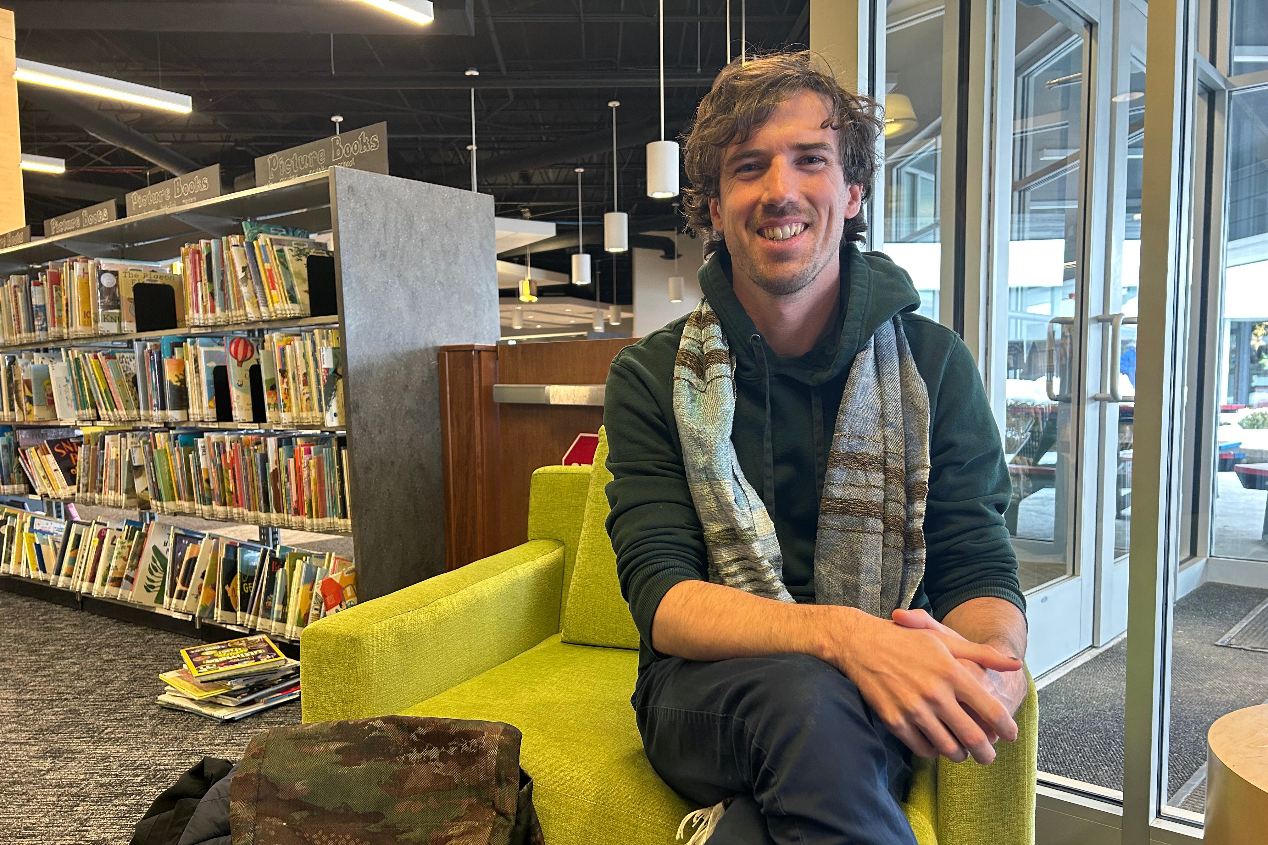 A man wearing a scarf sits on the side of a green chair and smiles at the camera with a bookshelf in the background.