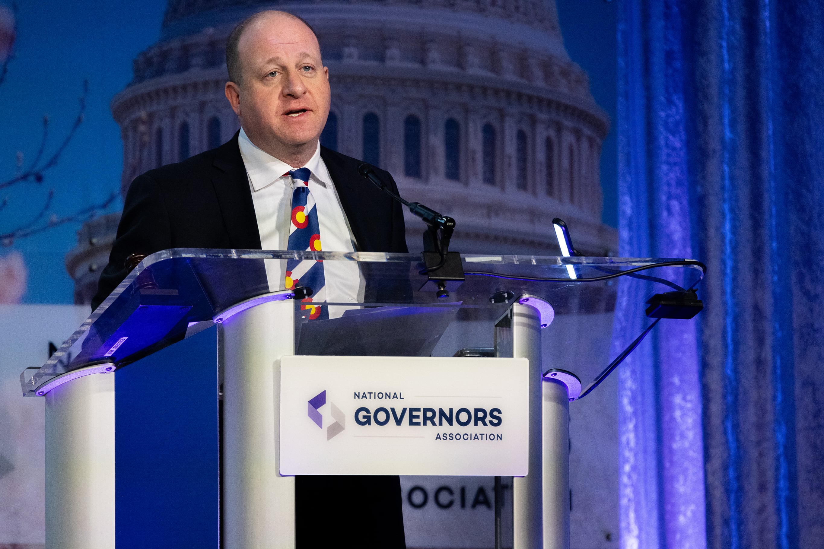 A photograph of a white man in a suit standing behind a podium on a stage.