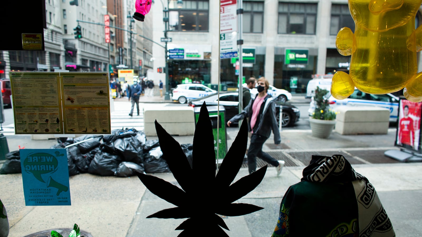 A marijuana plant is dark in the foreground in a store window while in the background people walk on the sidewalk outside.
