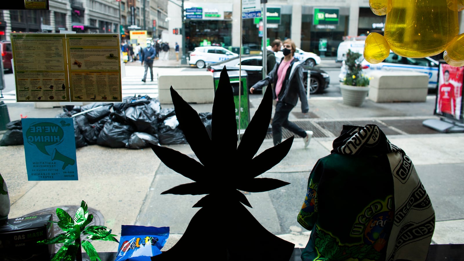 A marijuana plant is dark in the foreground in a store window while in the background people walk on the sidewalk outside.